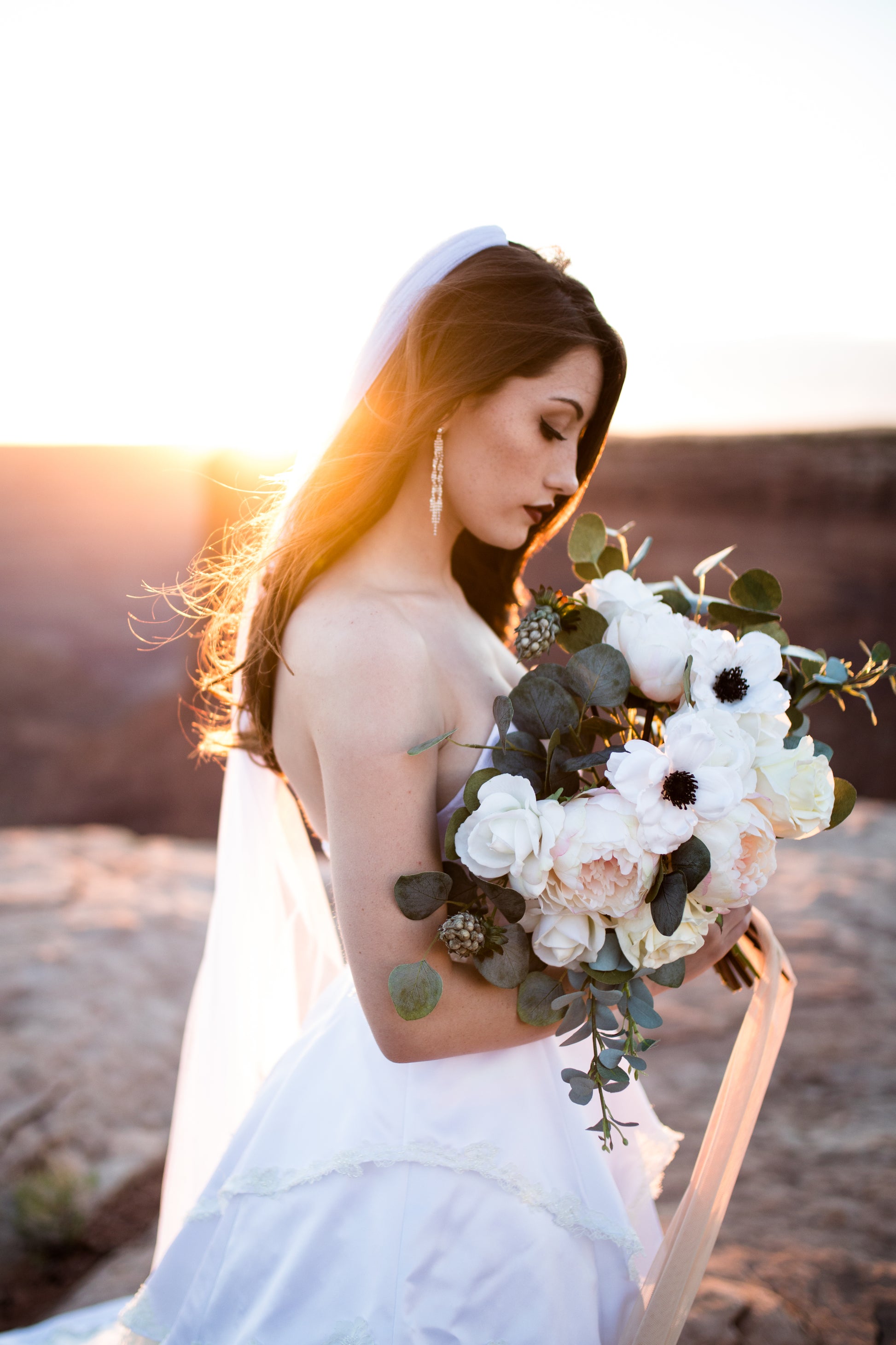 A photo of a bride at sunset; she stands to the side and holds a bridal boquet up. She is wearing a ballgown and the soft tulle jane veil. The veil is swept behind and to the side of the bride, caught in the breeze.