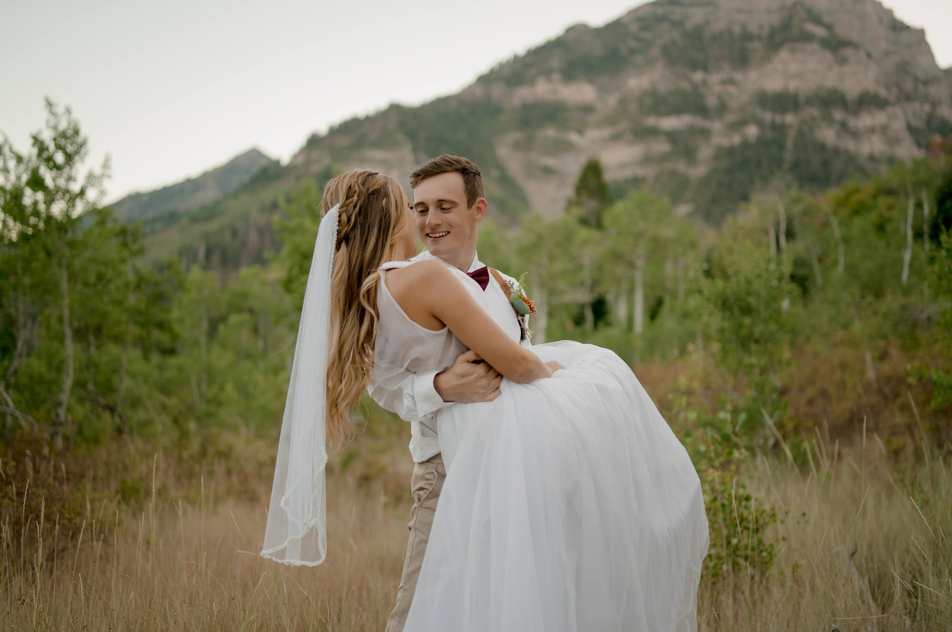 A groom holds his bride in a cradle lift with her back to camera so we can see the bridal veil as it falls from her hair. She wears the Lena veil with beaded trim edges and the tulle ballgown, Persephone.