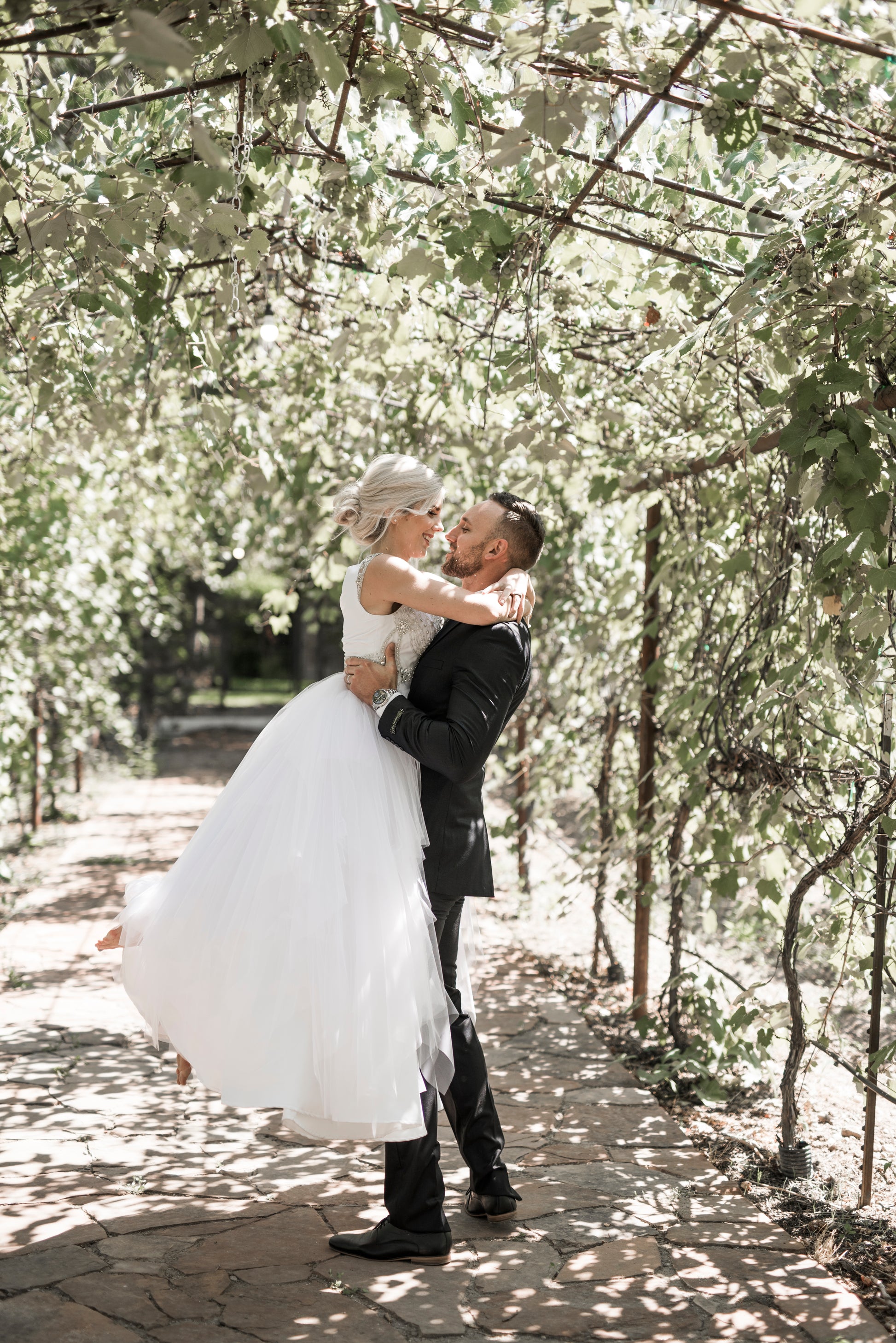 Groom picking up the bride while standing in a greenery arch. The side profile of the georgia gown is shown, showing off the skirt fullness and skirt tiers.