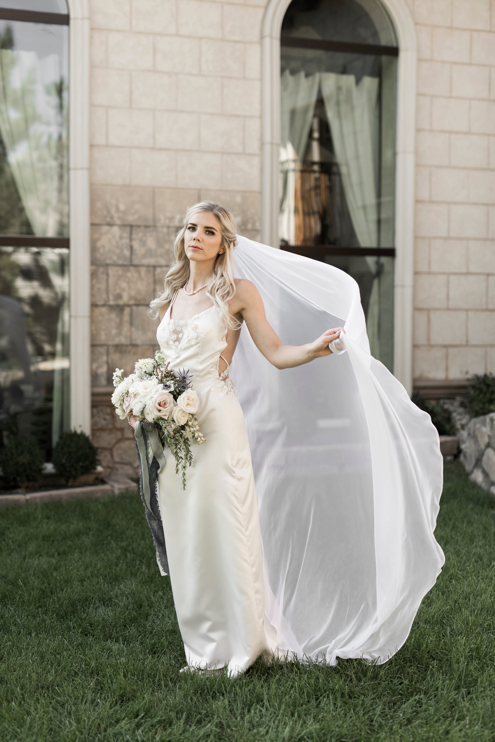 A bride wears the Braelyn veil in puddle length flowing behind her in outdoor ceremony. The veil has been caught in the wind and flows backward.
