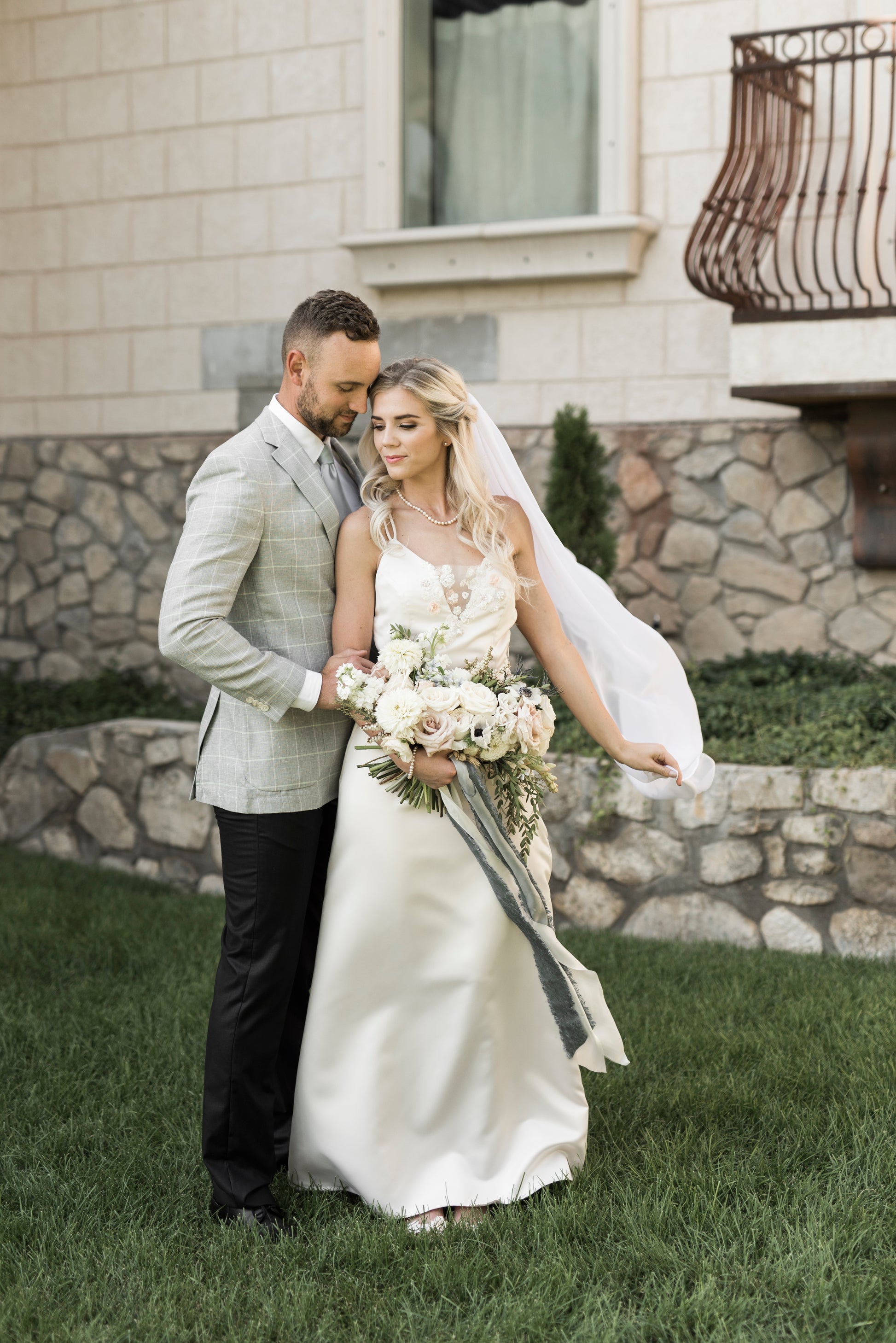 A photo of a bride and groom standing her back to his front; she wears the chiffon bridal veil as it gets caught in the wind to the side of her.