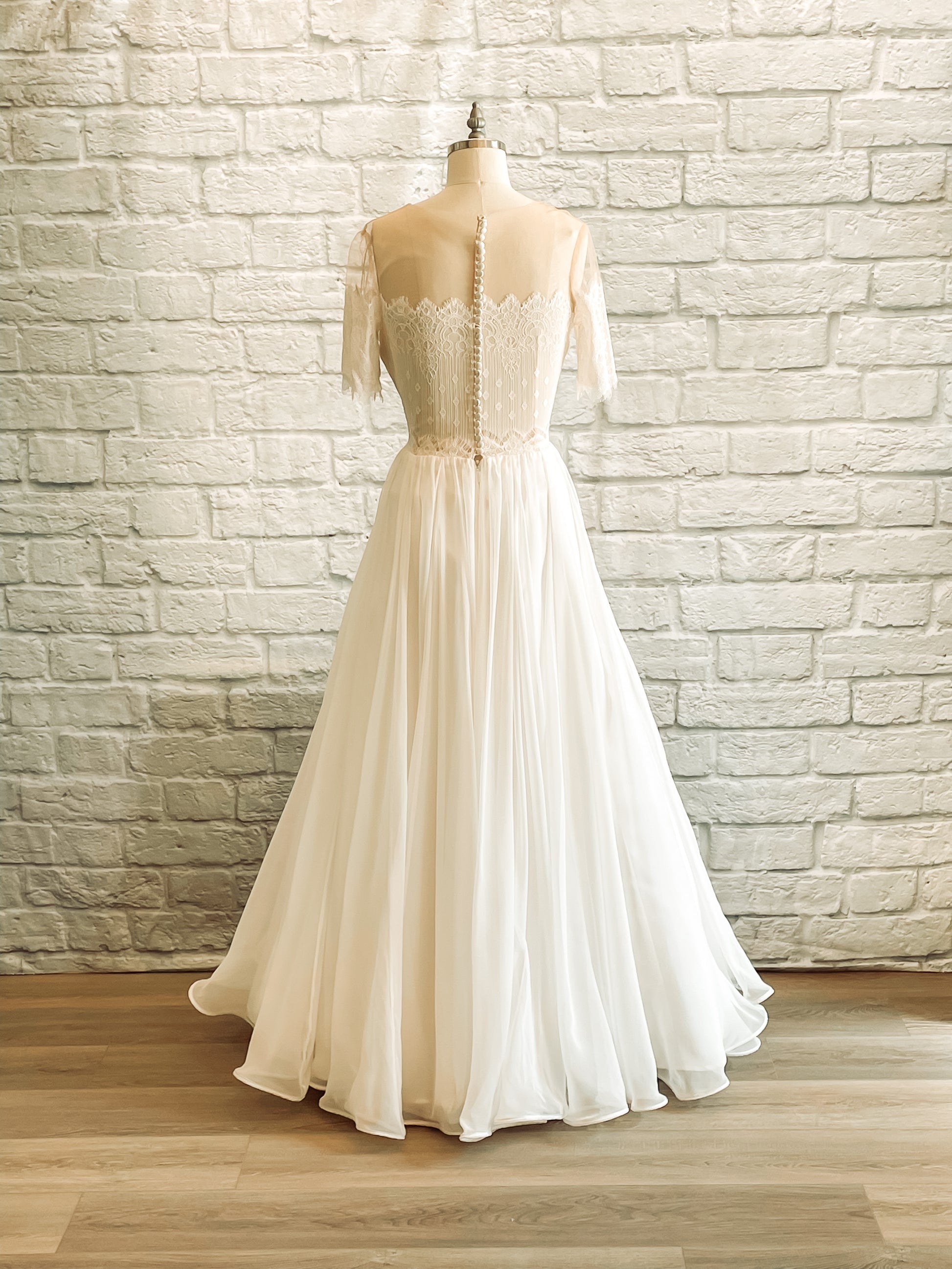 Full back view of the Ronelle wedding dress on a mannequin; this gown has a Sheer illusion back, satin buttons with a high scoop neckline, and a chiffon ball gown skirt.