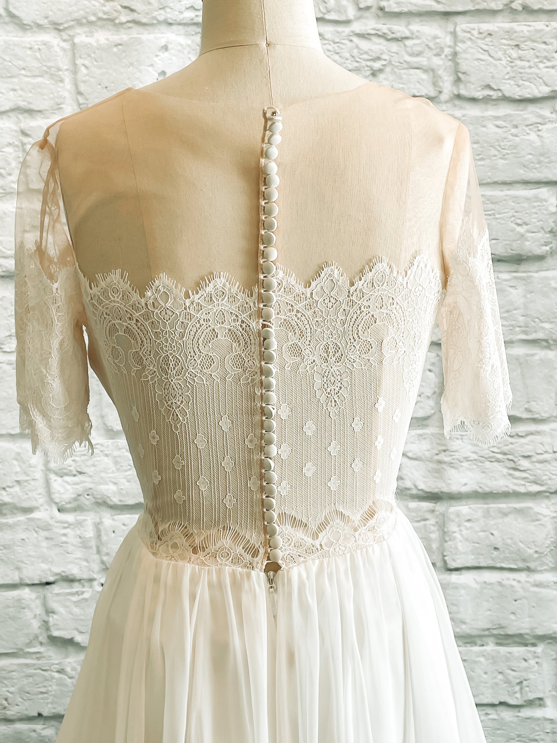 A close up shot of the Ronelle wedding dress and it's back bodie on a mannequin;  the gown has a sheer back that is closed with satin covered buttons.