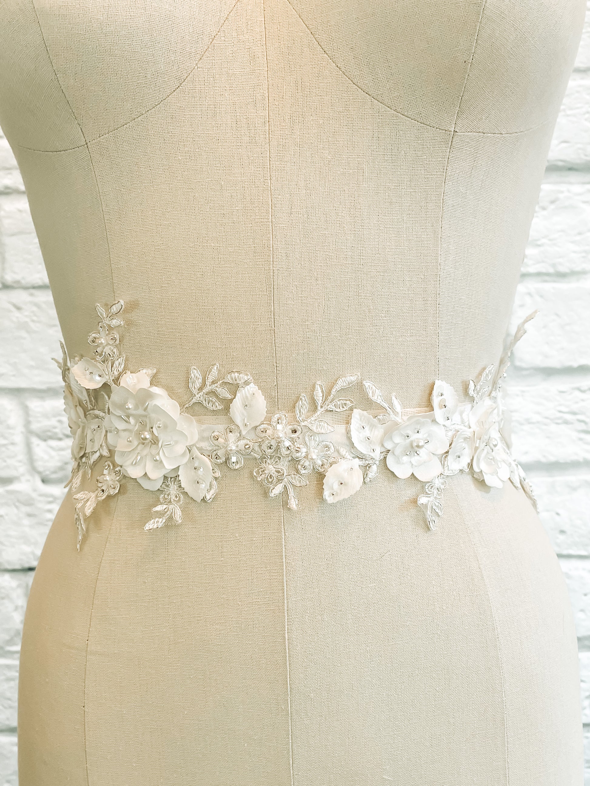 A front shot of the Meghan bridal belt on a mannequin; Close-up of Meghan bridal belt featuring satin 3D flowers, pearl accents, and metallic thread on a satin ribbon.