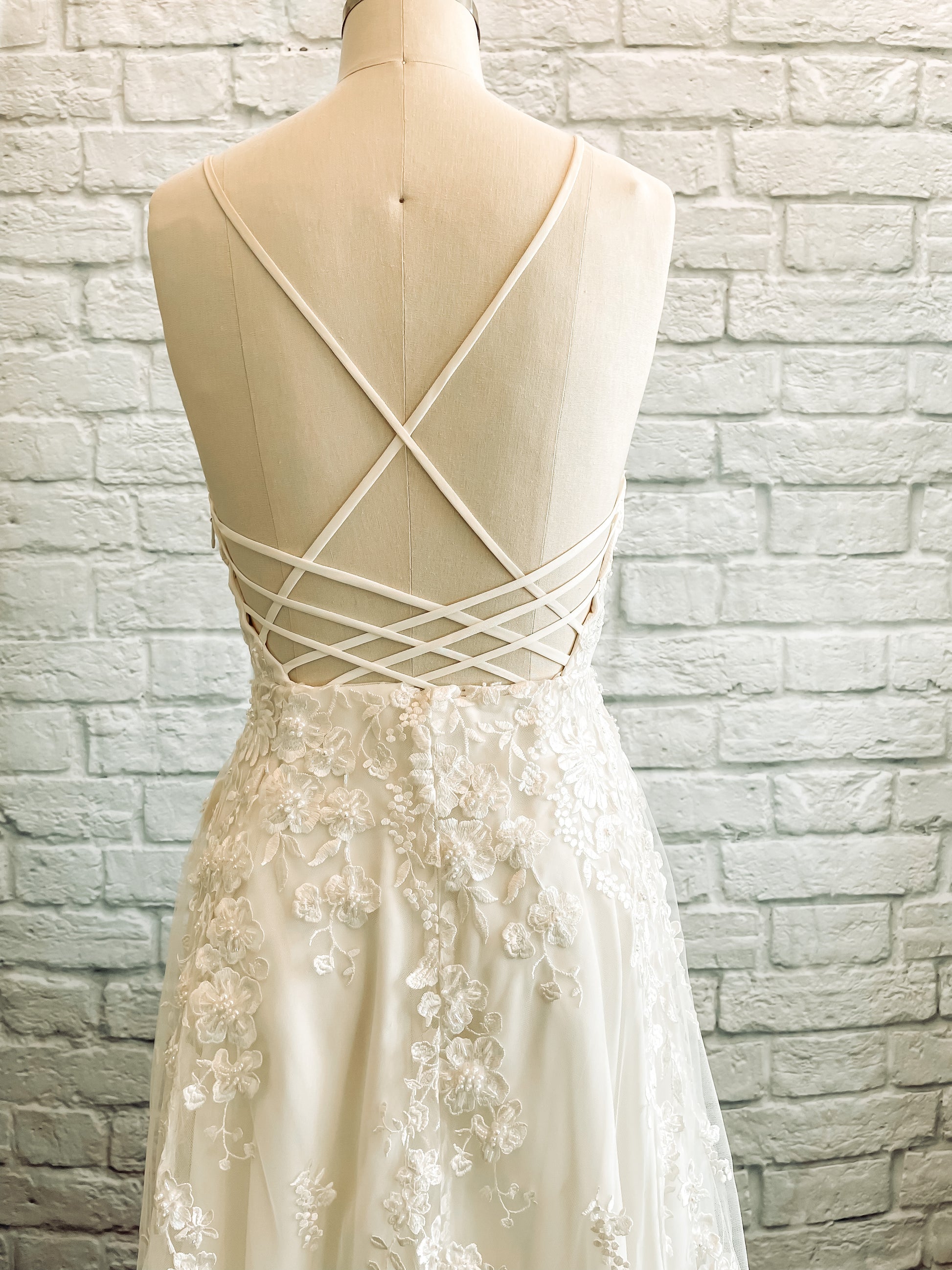 A close up of the back bodice and top of the skirt of the Wilhelmina gown on a mannequin showing off the criss cross straps and Detailed floral lace with pearl and bead embellishments