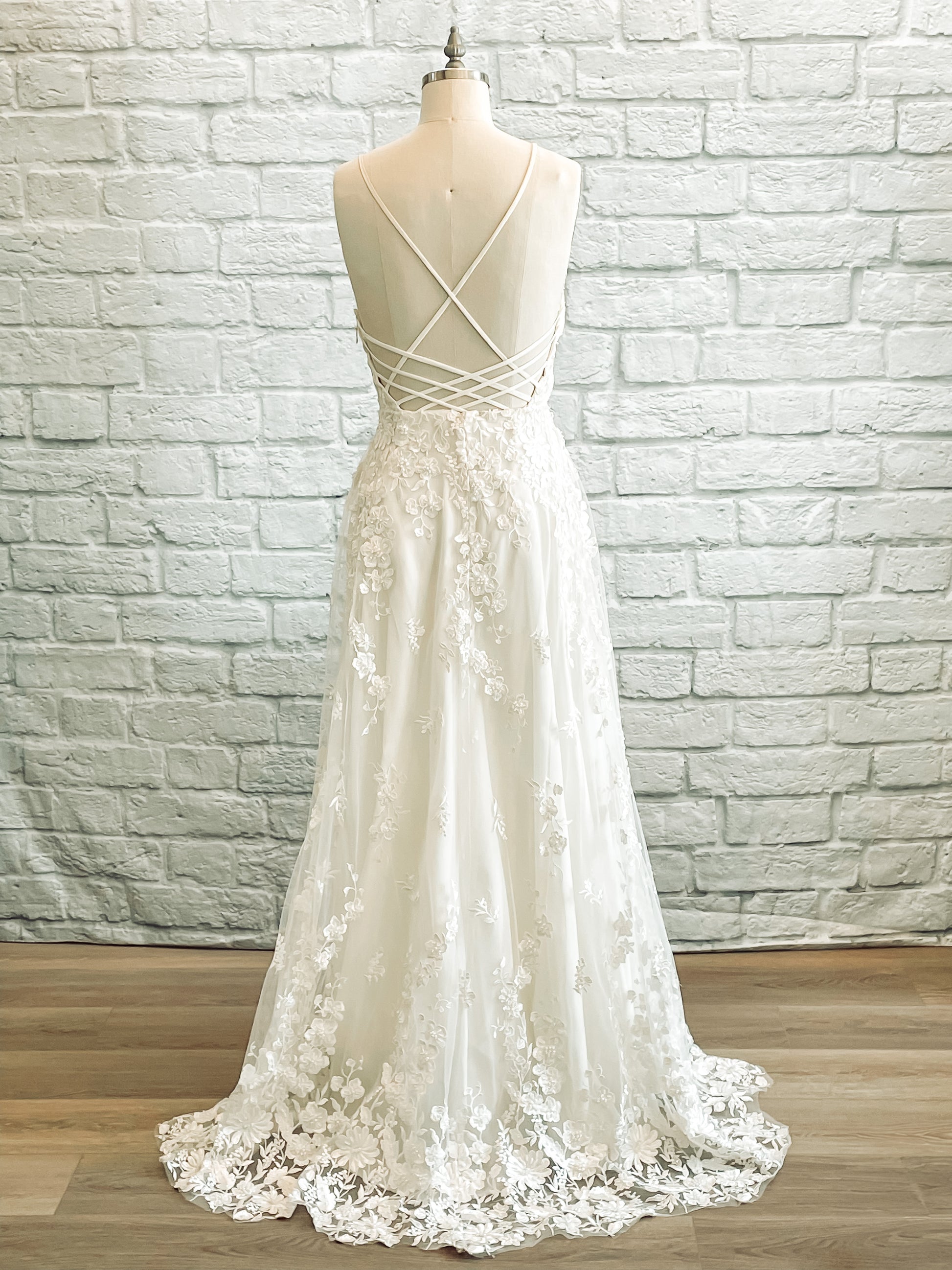 A full length shot of the wilhelmina gown from the back; the gown has  Crisscrossing straps over an open back, lace skirt, and small lace train detail