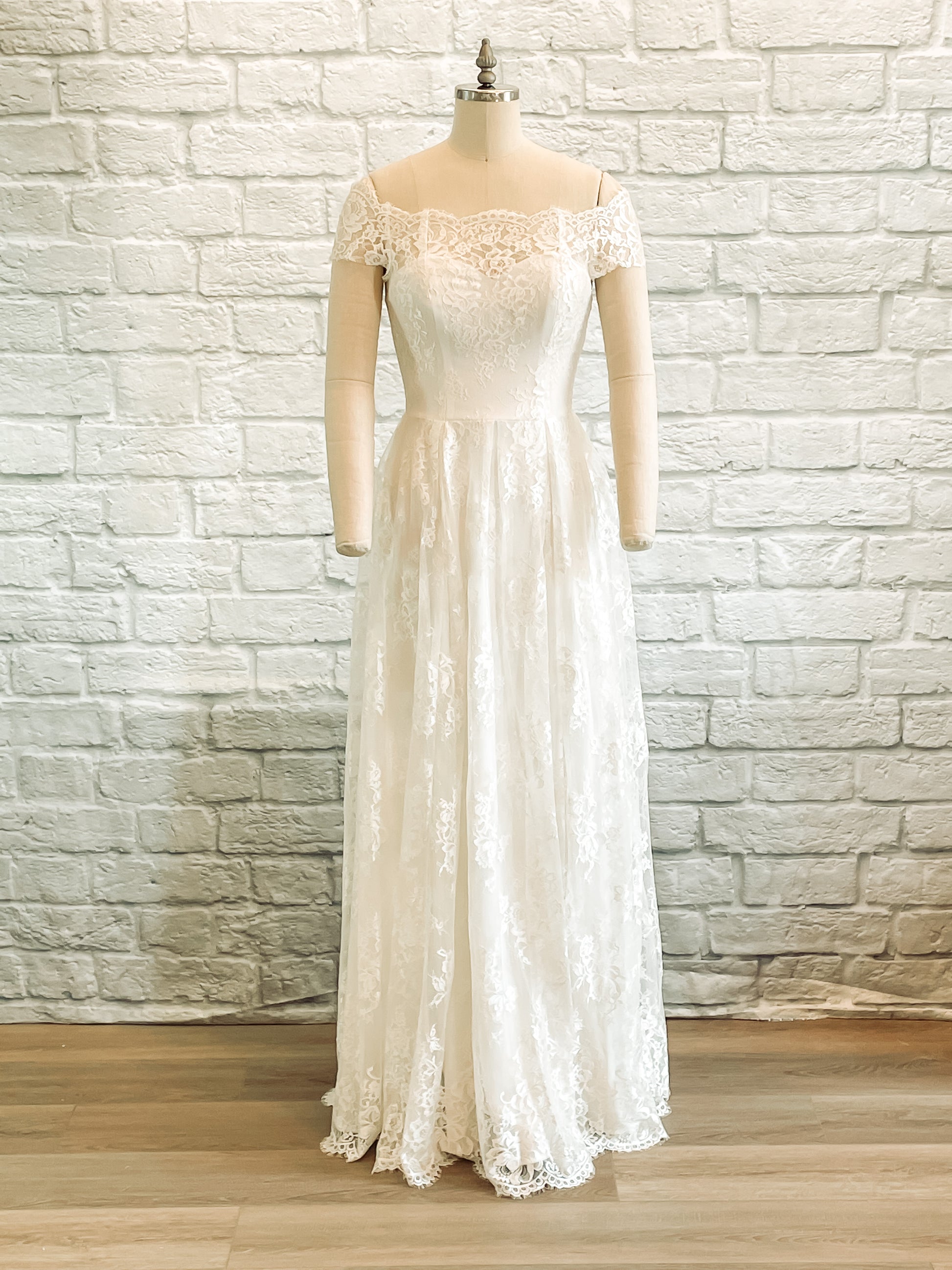A full length photo of the Serafina gown on a mannequin showing the front of the dress, the sweetheart neckline, box pleated skirt, and off-the-shoulder lace sleeves are all shown off.