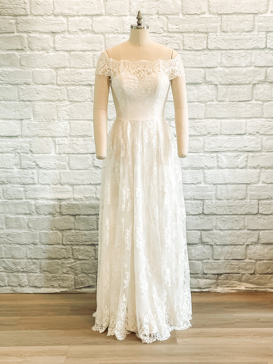 A full length photo of the Serafina gown on a mannequin showing the front of the dress, the sweetheart neckline, box pleated skirt, and off-the-shoulder lace sleeves are all shown off.