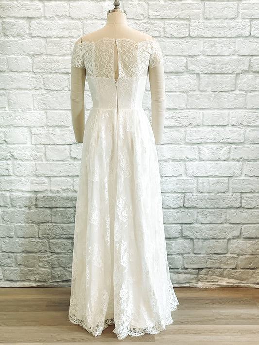 A full length photo of the Serafina gown on a mannequin showing the back of the dress and the straight-across crepe back with the off-the-shoulder lace sleeve structure.
