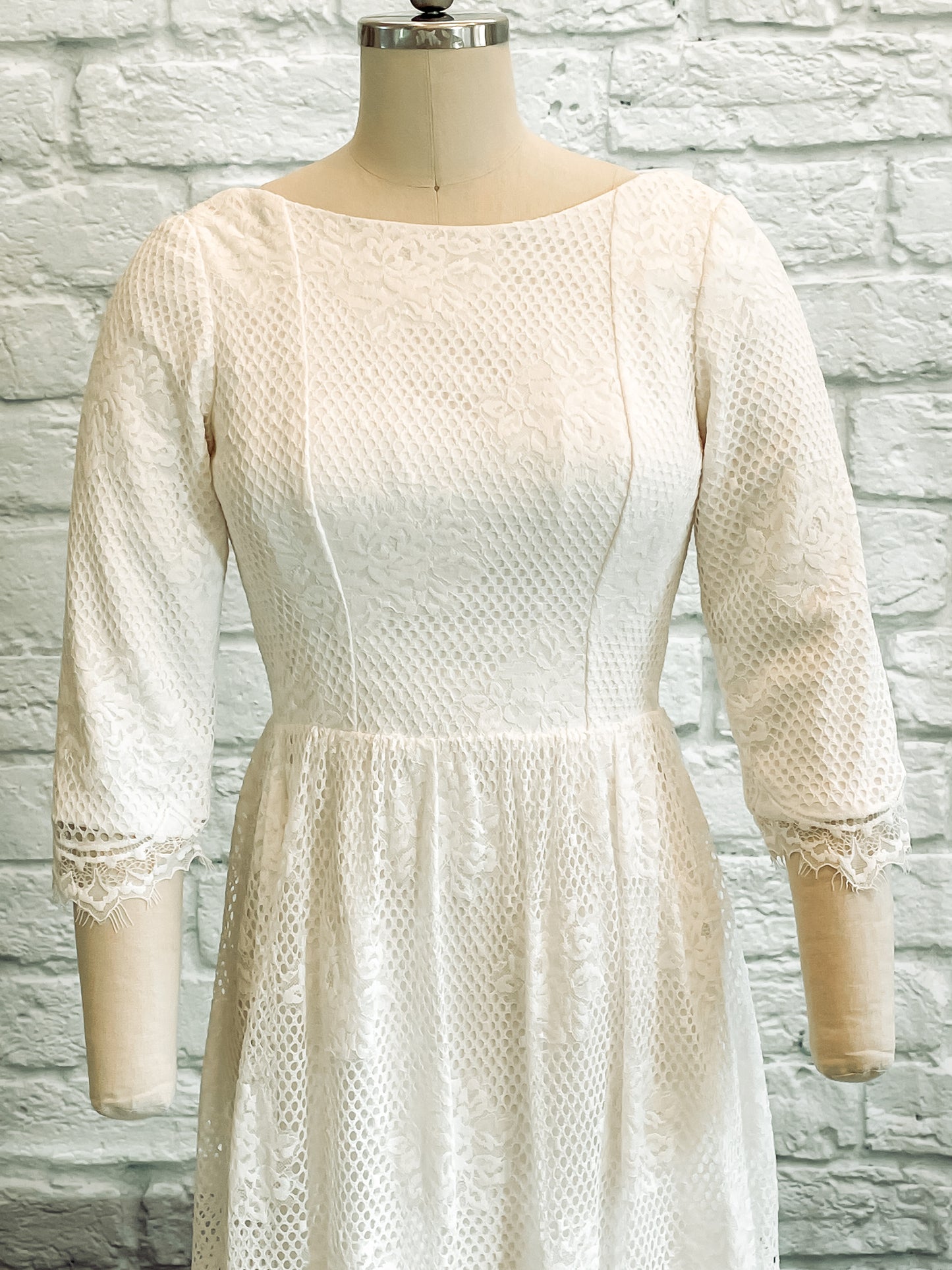 Detail shot of Avabelle gown’s front bodice , lace sleeves and wide boat neckline on a mannequin, highlighting the rose lace pattern and scalloped eyelash edges.