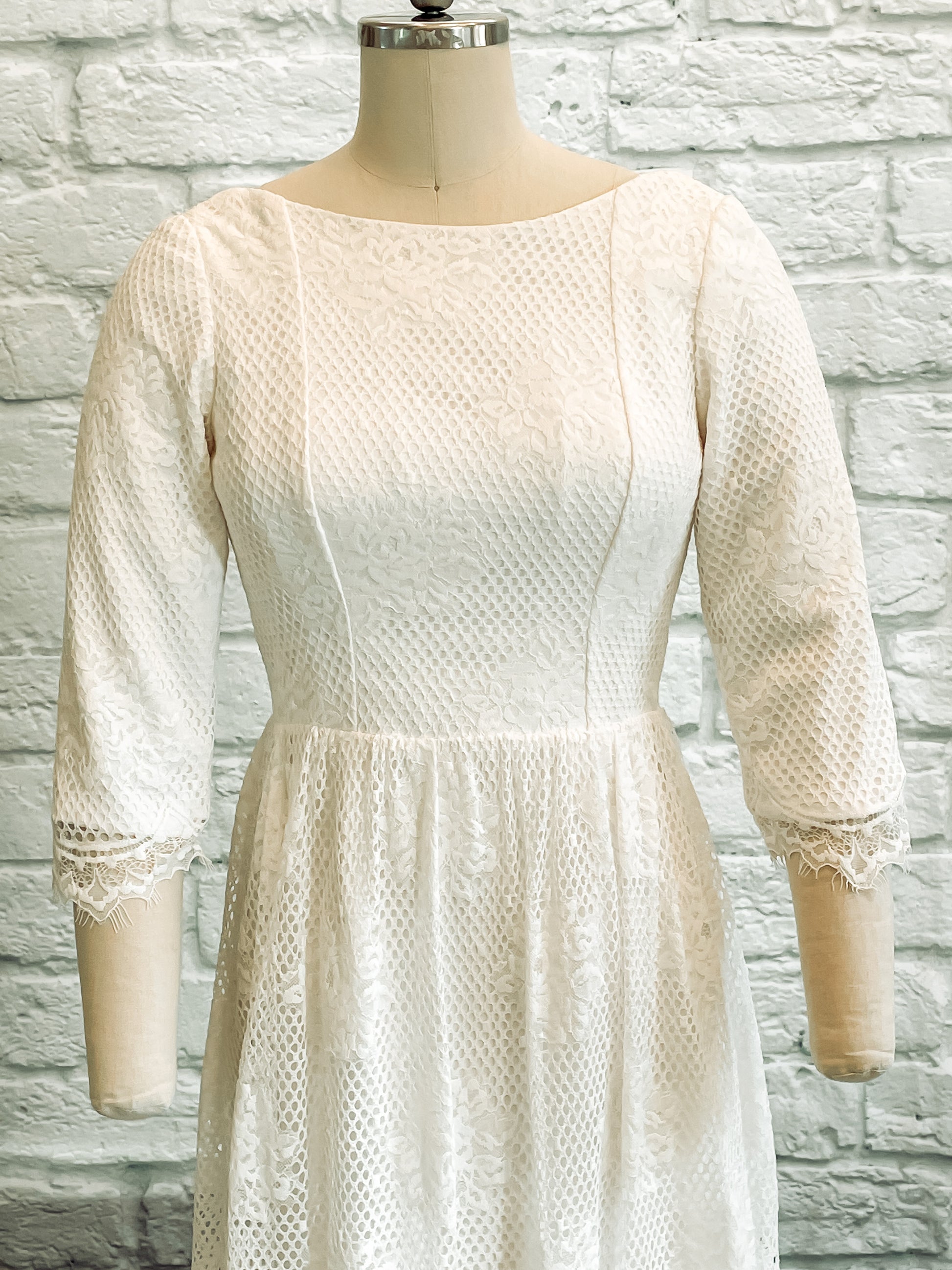 Detail shot of Avabelle gown’s front bodice , lace sleeves and wide boat neckline on a mannequin, highlighting the rose lace pattern and scalloped eyelash edges.