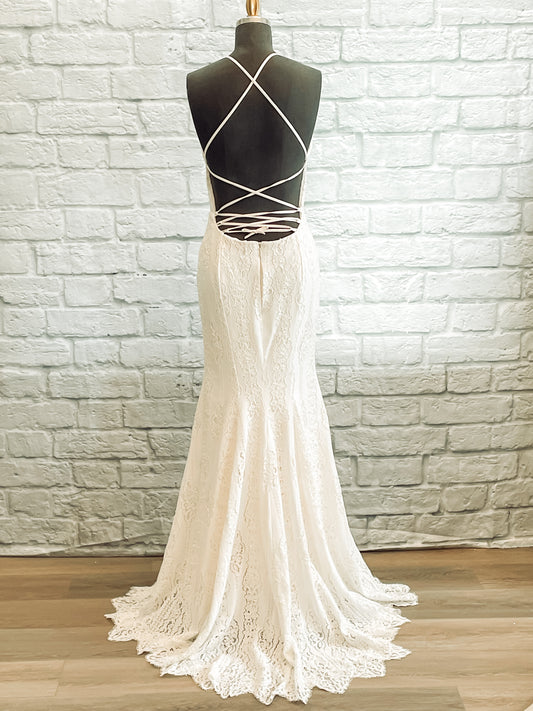 A full length back image of the Willoughby gown on a mannequin; the gown has crisscross spaghetti straps between the deep scoop back and a soft small train with irregularly shaped scallop.
