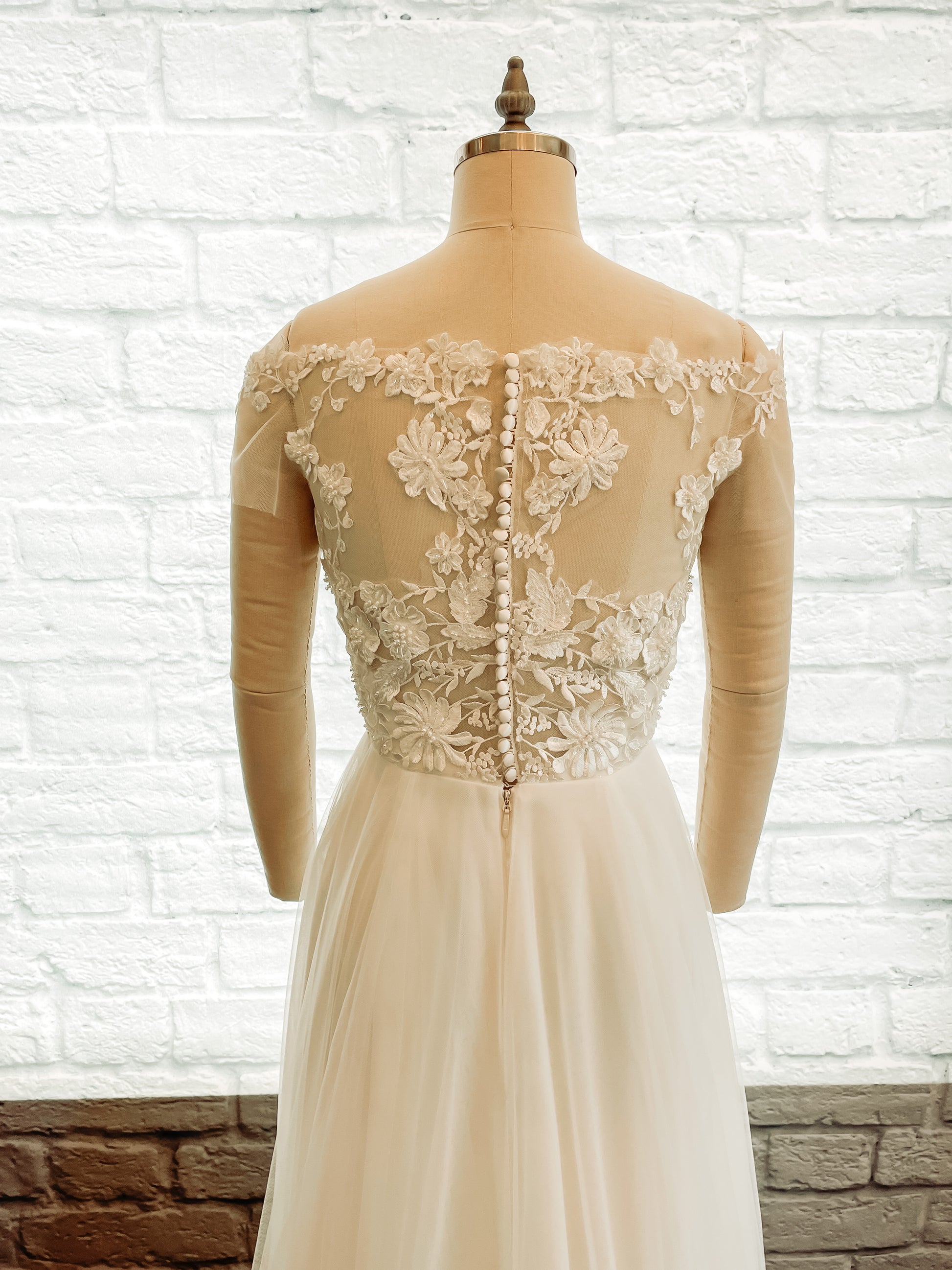 Up close of the gowns back bodice on a mannequin, showing the soft bardot neckline, off-shoulder tulle sleeves and satin covered bridal buttons.