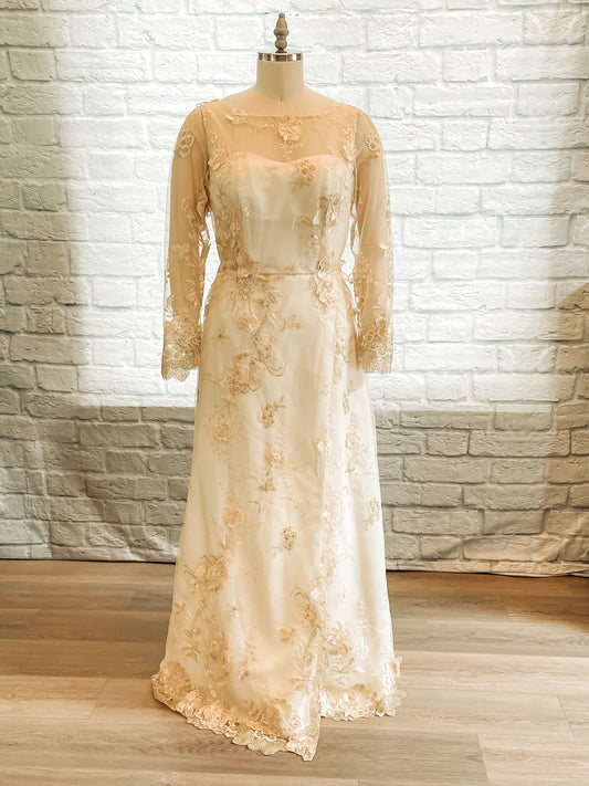 A full front view of the Reagan wedding gown with gold lace overlay, long lace sleeves, and mock wrap skirt.