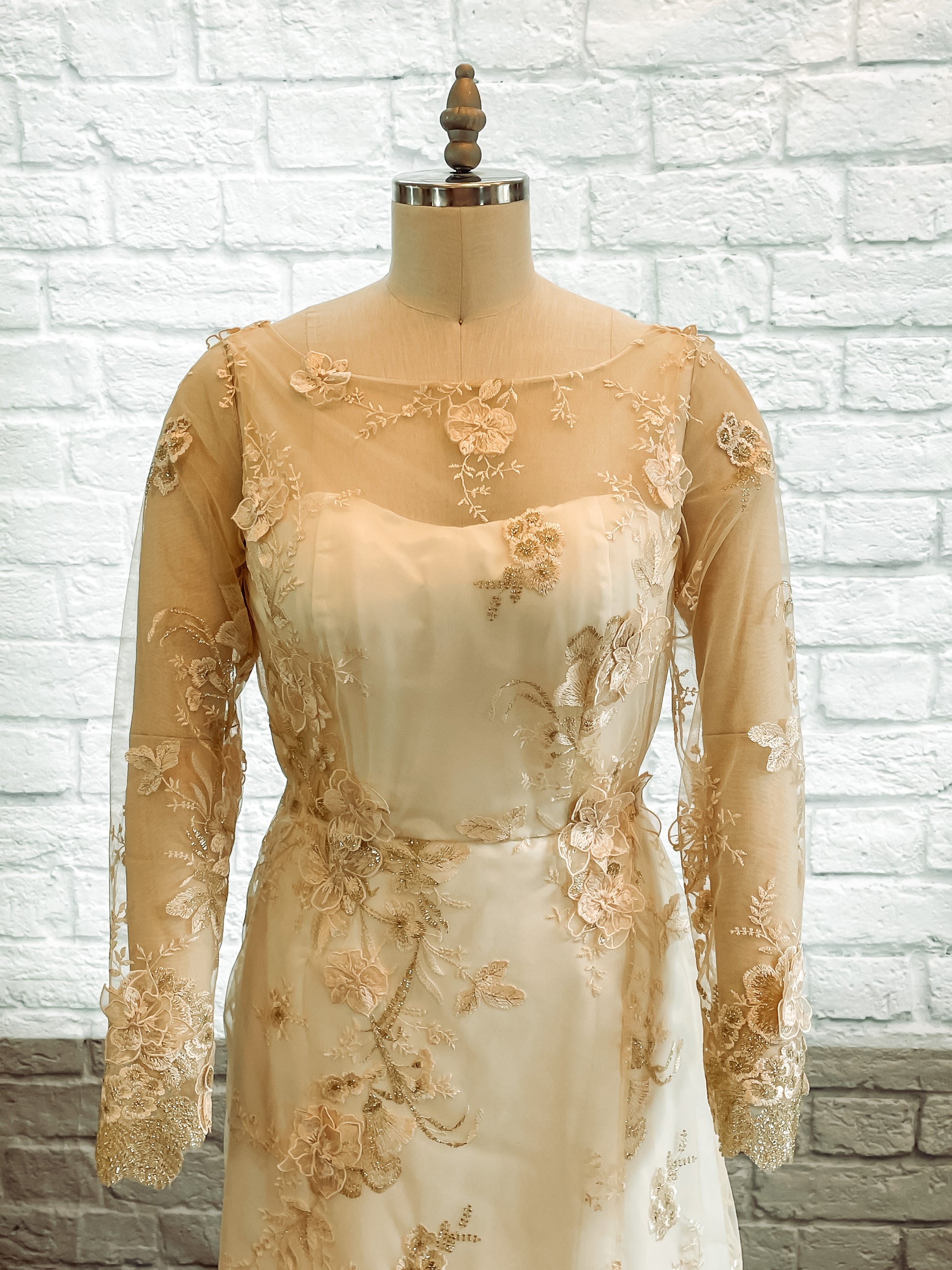 A close up image of the Reagan gown on a mannequin  showing off the Boat neckline with sheer gold floral lace over sweetheart base and long lace sleeves.
