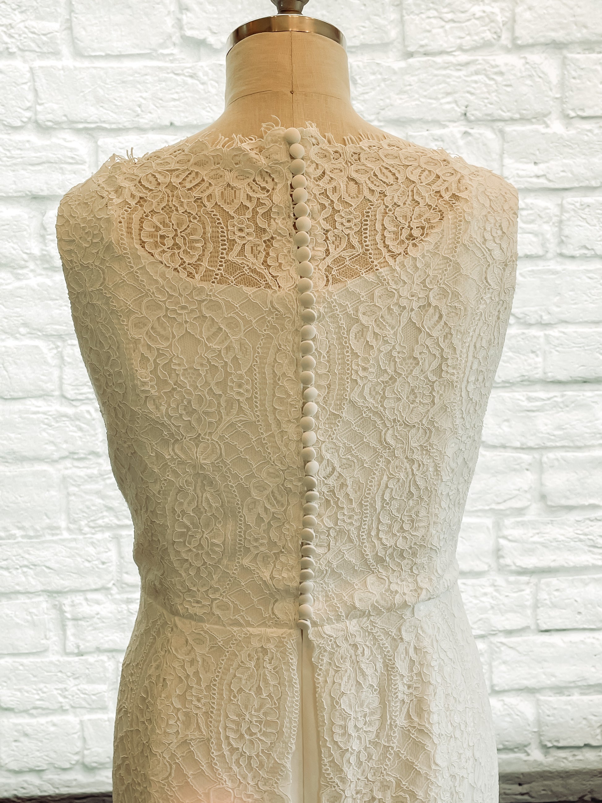 Close-up of the back bodice of the Blair gown on a mannequin, featuring satin-covered buttons running down the center of the lace overlay, with a wide scalloped boat neckline and intricate floral lace detailing.