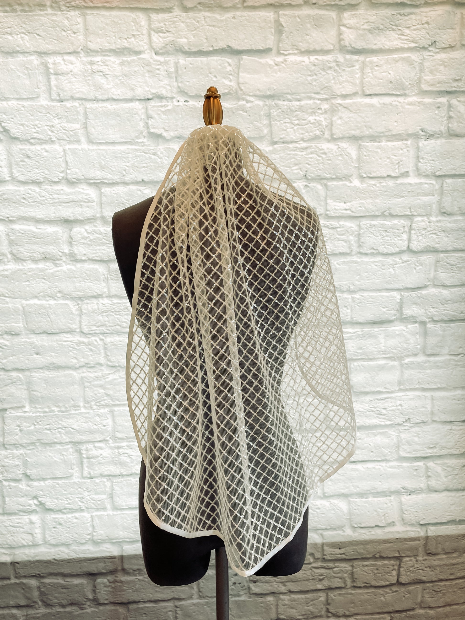 The gloria veil has been displayed on a mannequin with the veil pulled over one of the shoulders and not the other. This veil is covered in sequins in a diamond pattern and ribbon trim edge creating a detailed fairytale look.