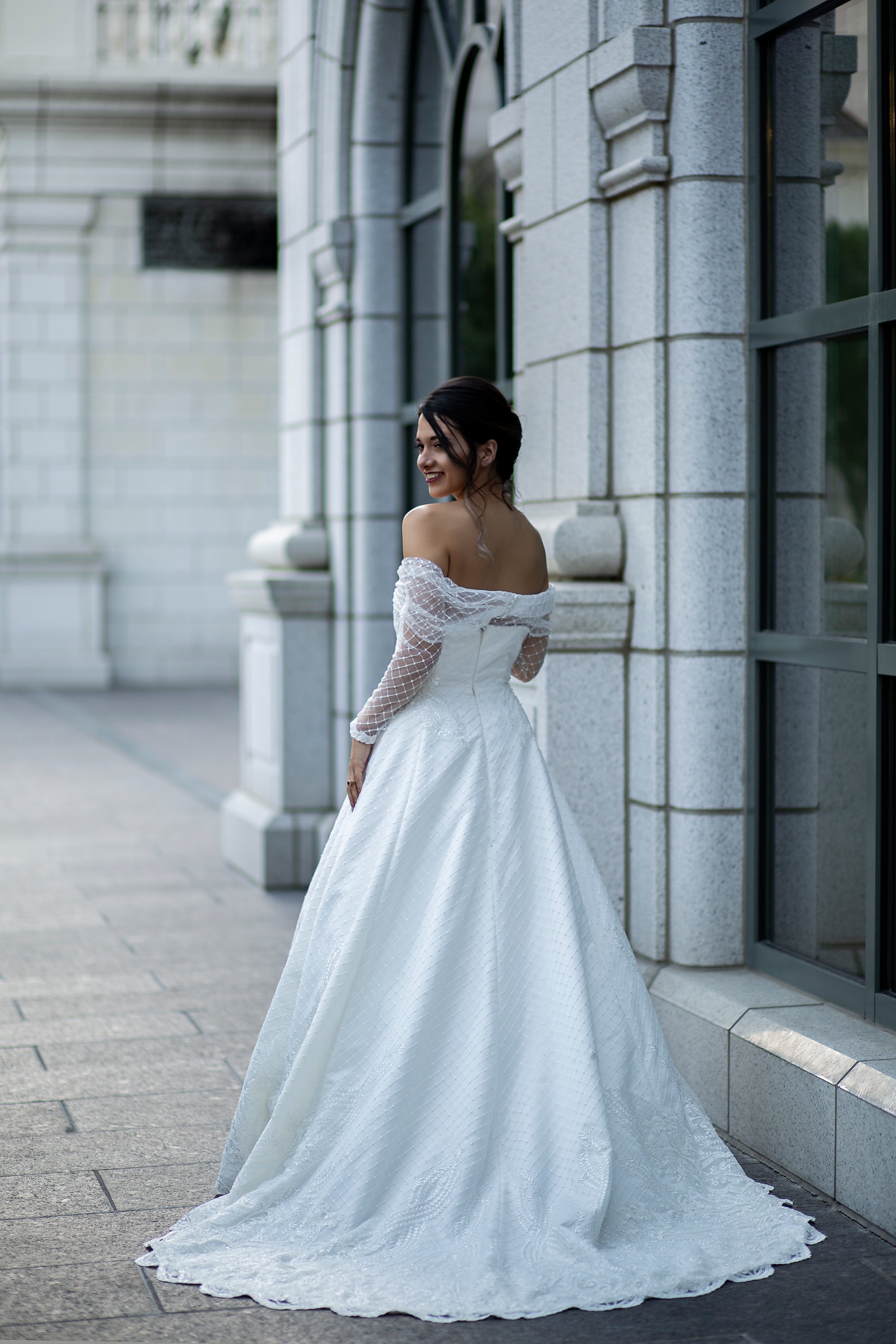 A full length view of the Virginia gown on a bride; the gowns train, off the shoulder bardot neckline and long sleeves are all shown off in this photo.