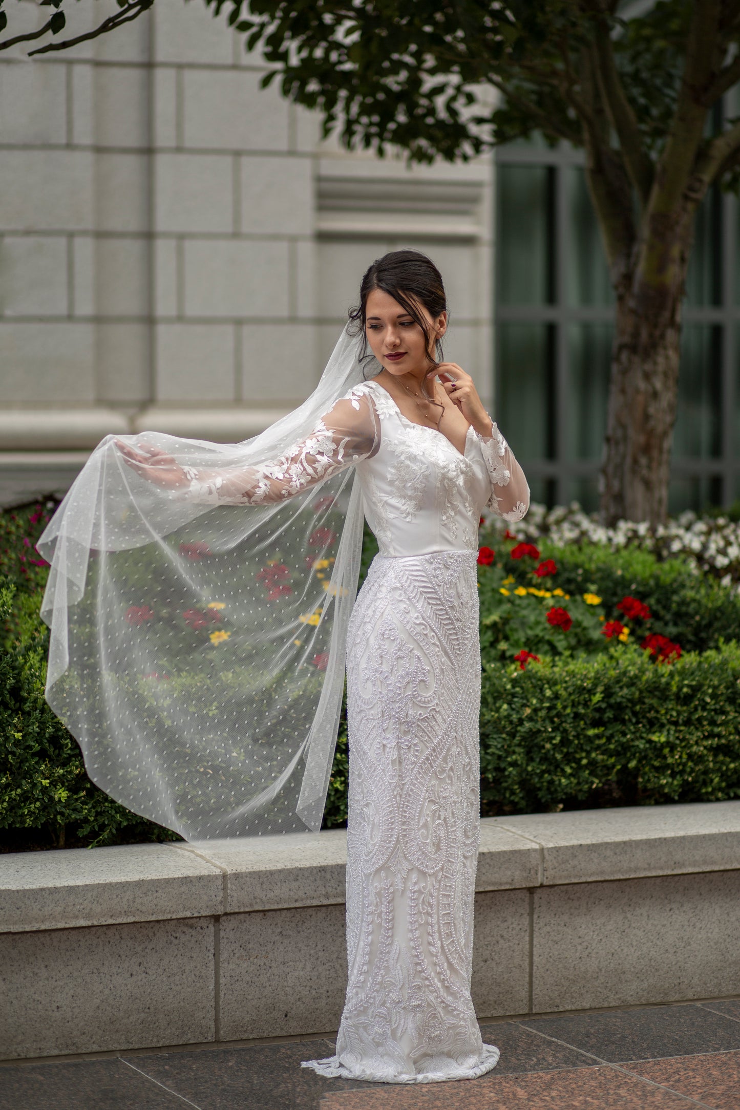 Bride in Swiss Dot veil, Scarlett, paired with the sheath lace gown, Octavia. The image is a full front of the bride standing almost ideways with her back arm outstretched and the veil reasting on top and then flowing down.