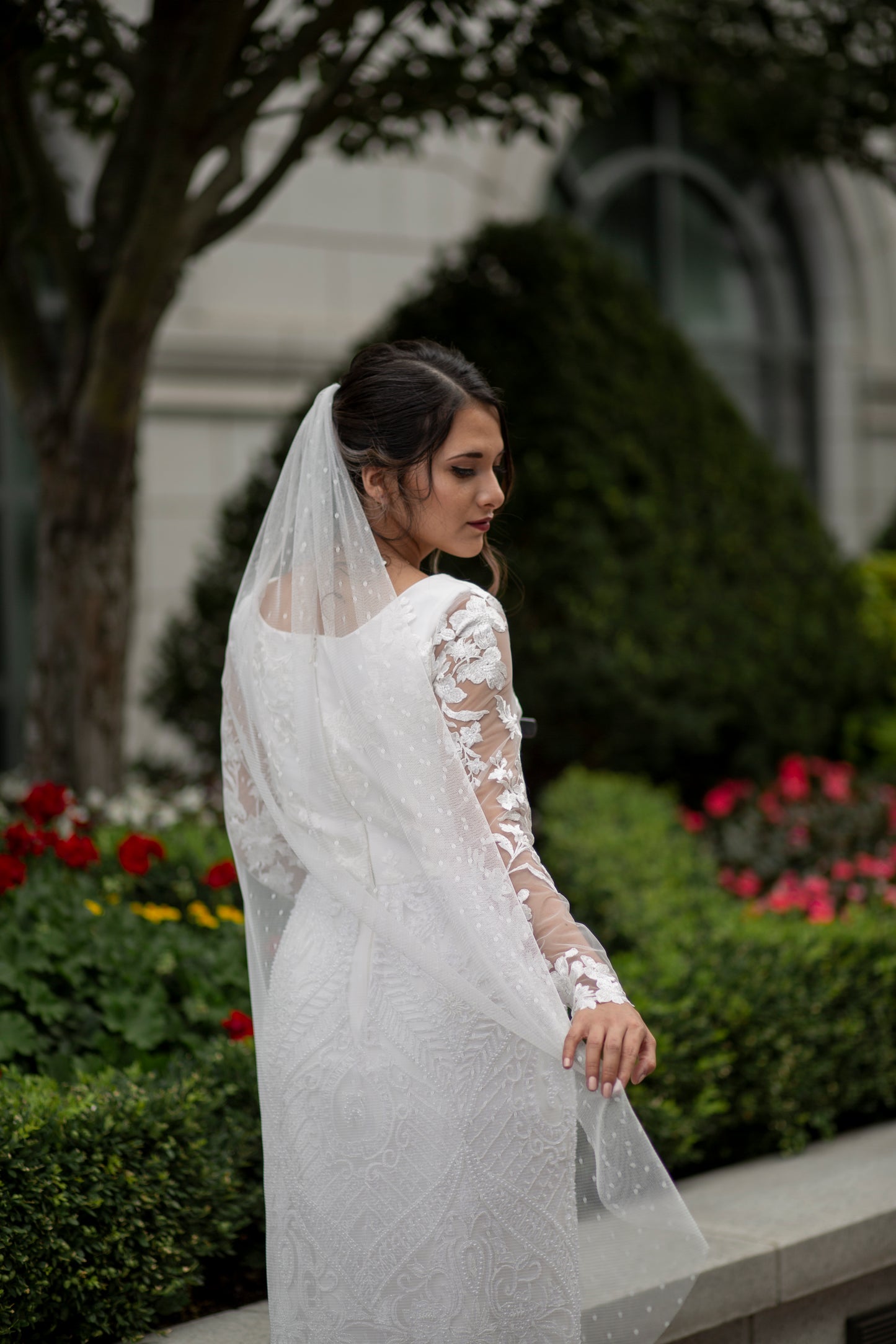 in this photo the scarlett veil is being paired with the octavia gown. a bride stands with her back to us and holds the edge of the swiss dot veil out behind her. The waltz-length veil with playful Swiss Dot texture pairs with the all lace gown octavia.
