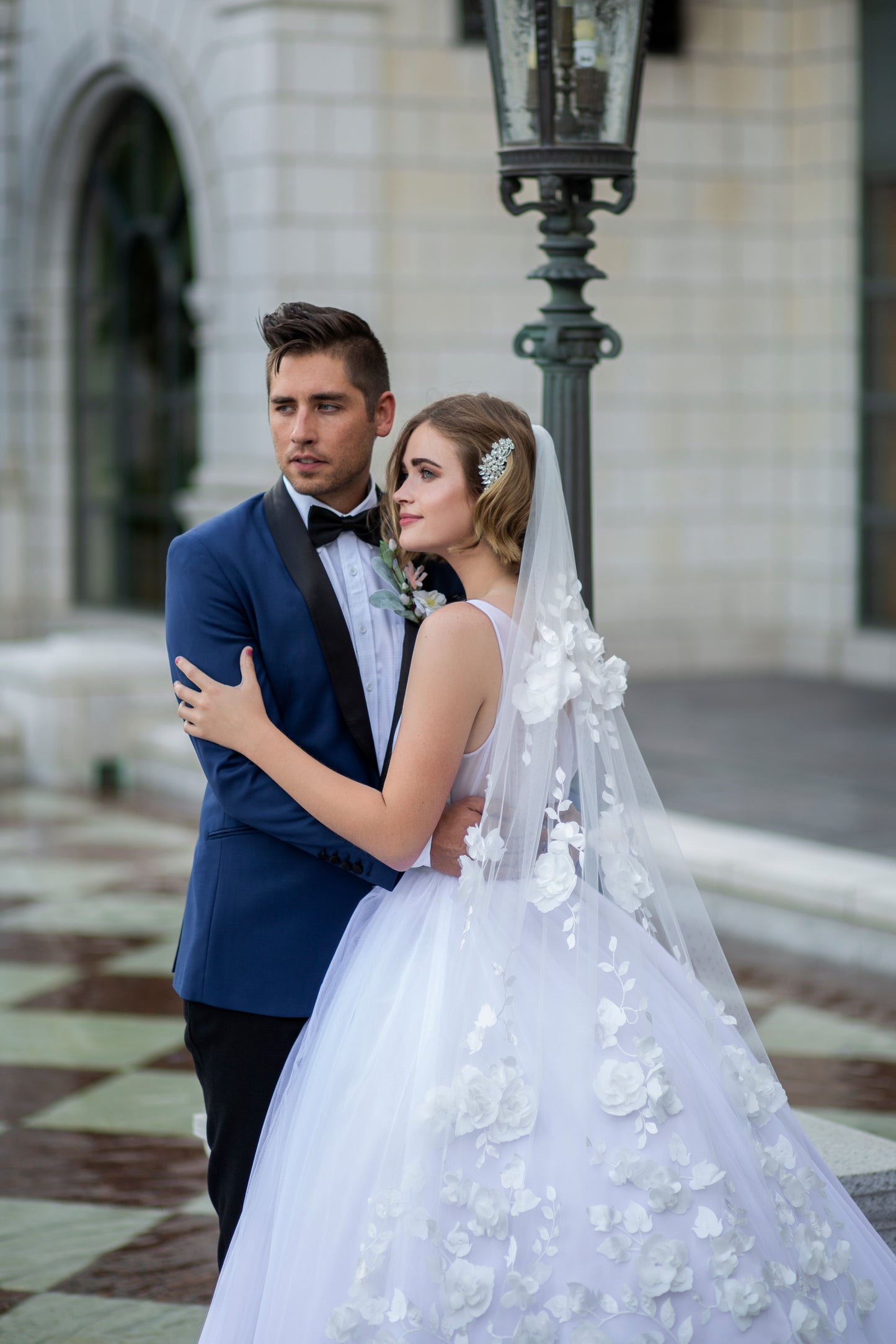 A bride and groom stand together embrasing. She wears the persephone gown and Celine veil. The veil is the focal point of the image as it lays over the ball gown showing off the 3d flowers.