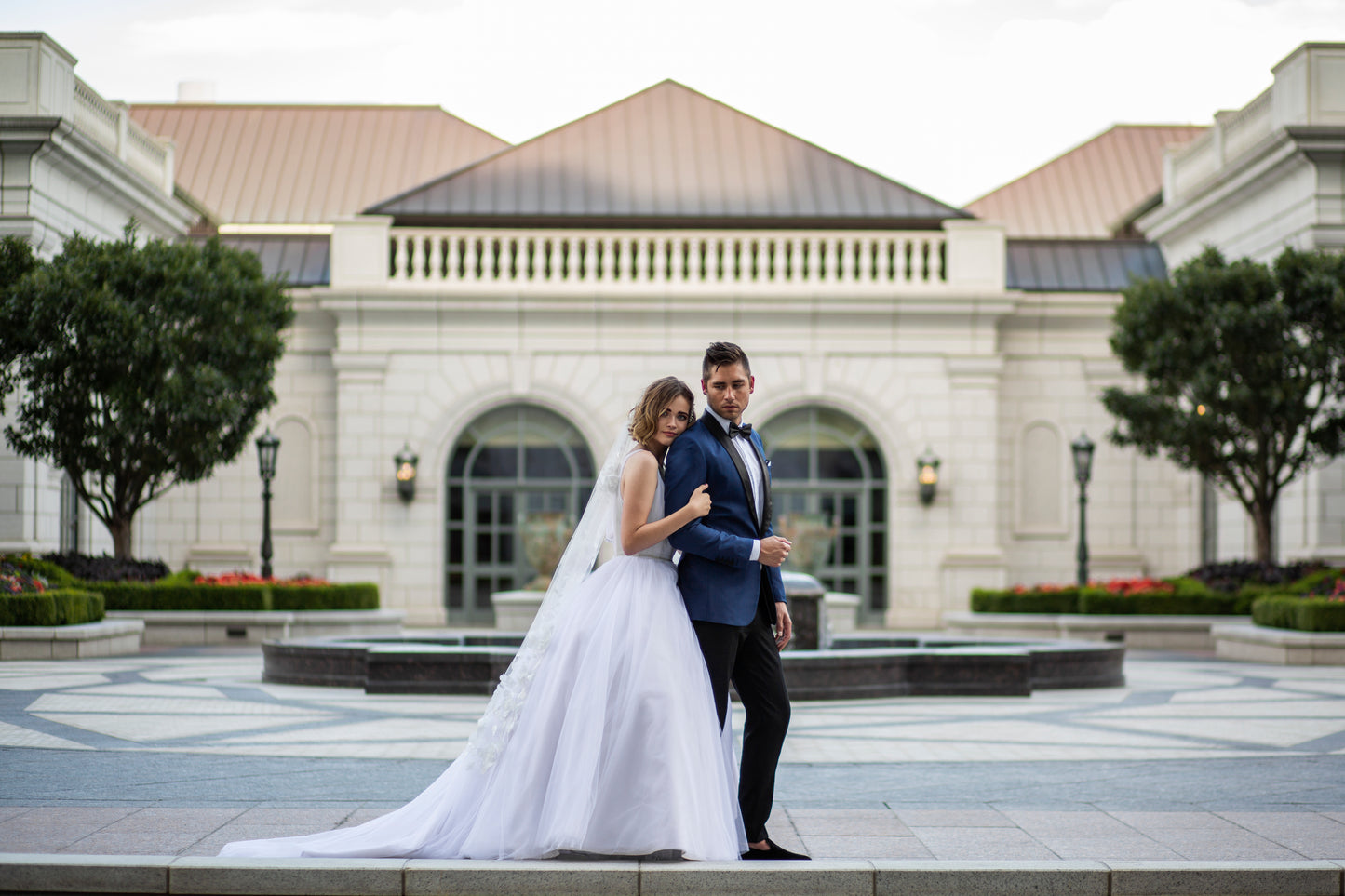 A bride leans against her husbands shoulder from behind and they both look at the camera from their profile. The bride wears the Persephone gown and the Celine veil; the veil is in pale ivory waltz length and has cascading 3d floral details.