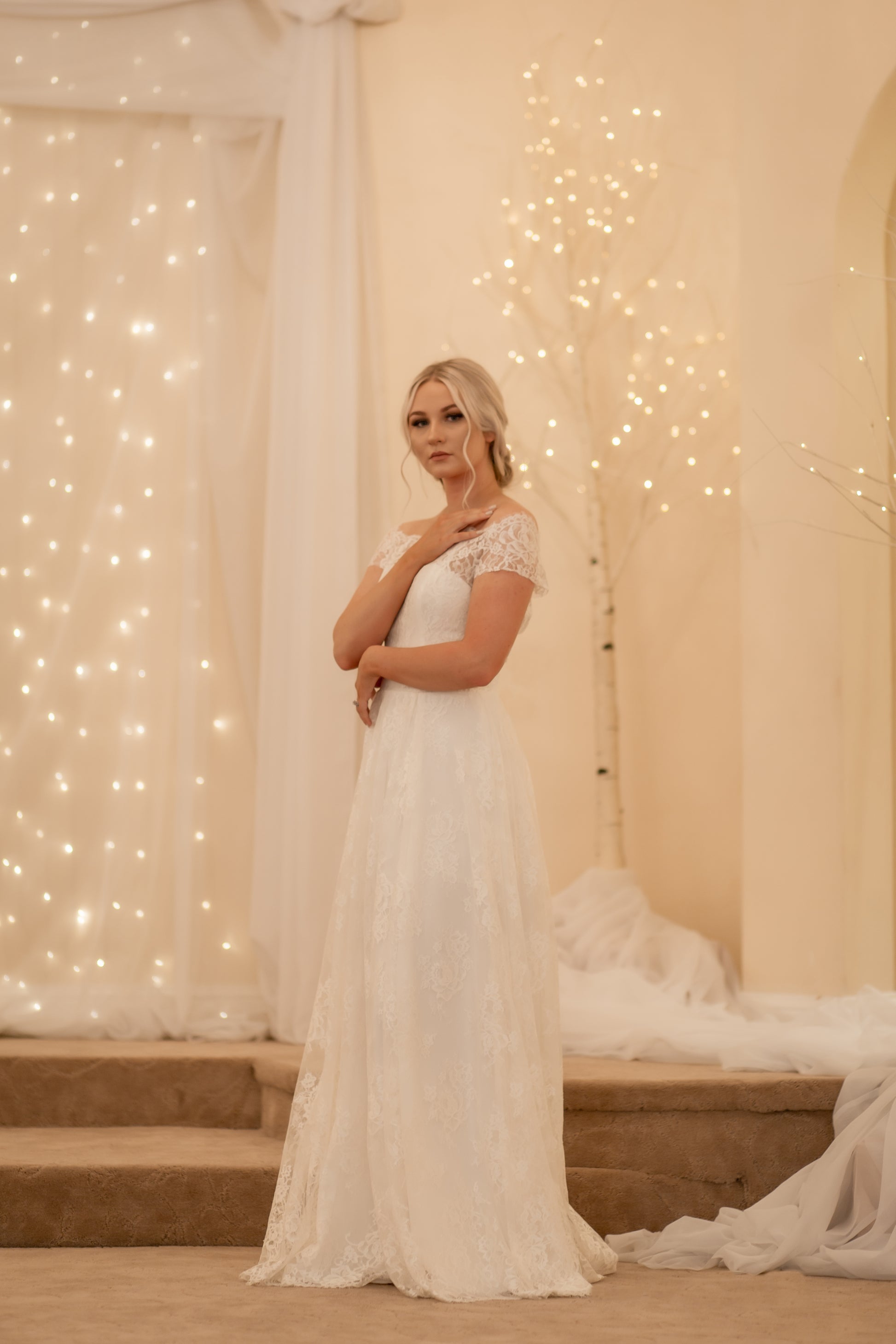 A side view of the Serafina gown; the bride wears it and looks at camera. The wedding gowns Modified A-line crepe base with lace overlay are on display.