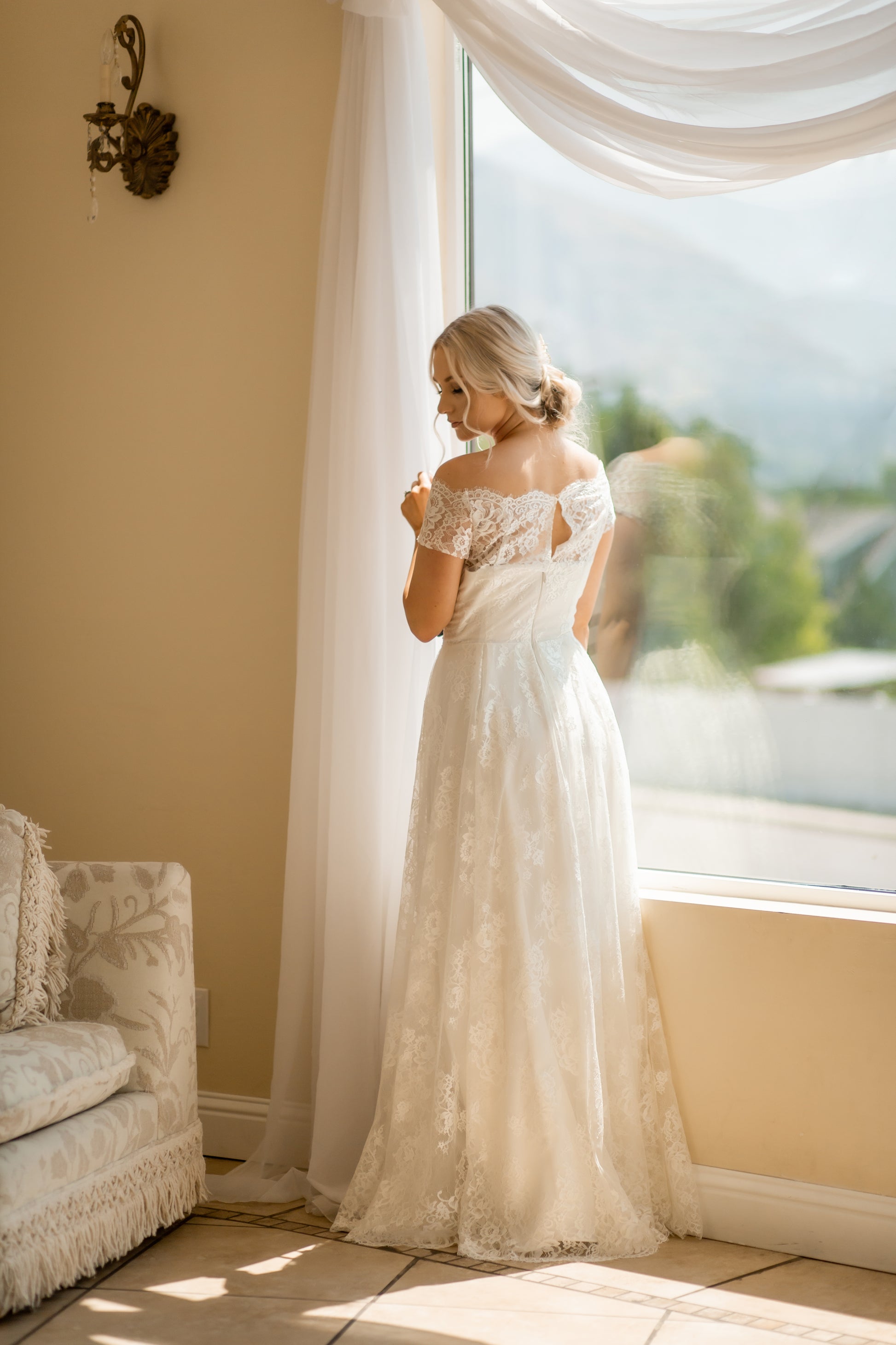 A full length photo of the back of Serafina; the a-line silhouette, off the shoulder lace sleeves, and the salloped neckline are all shown off.