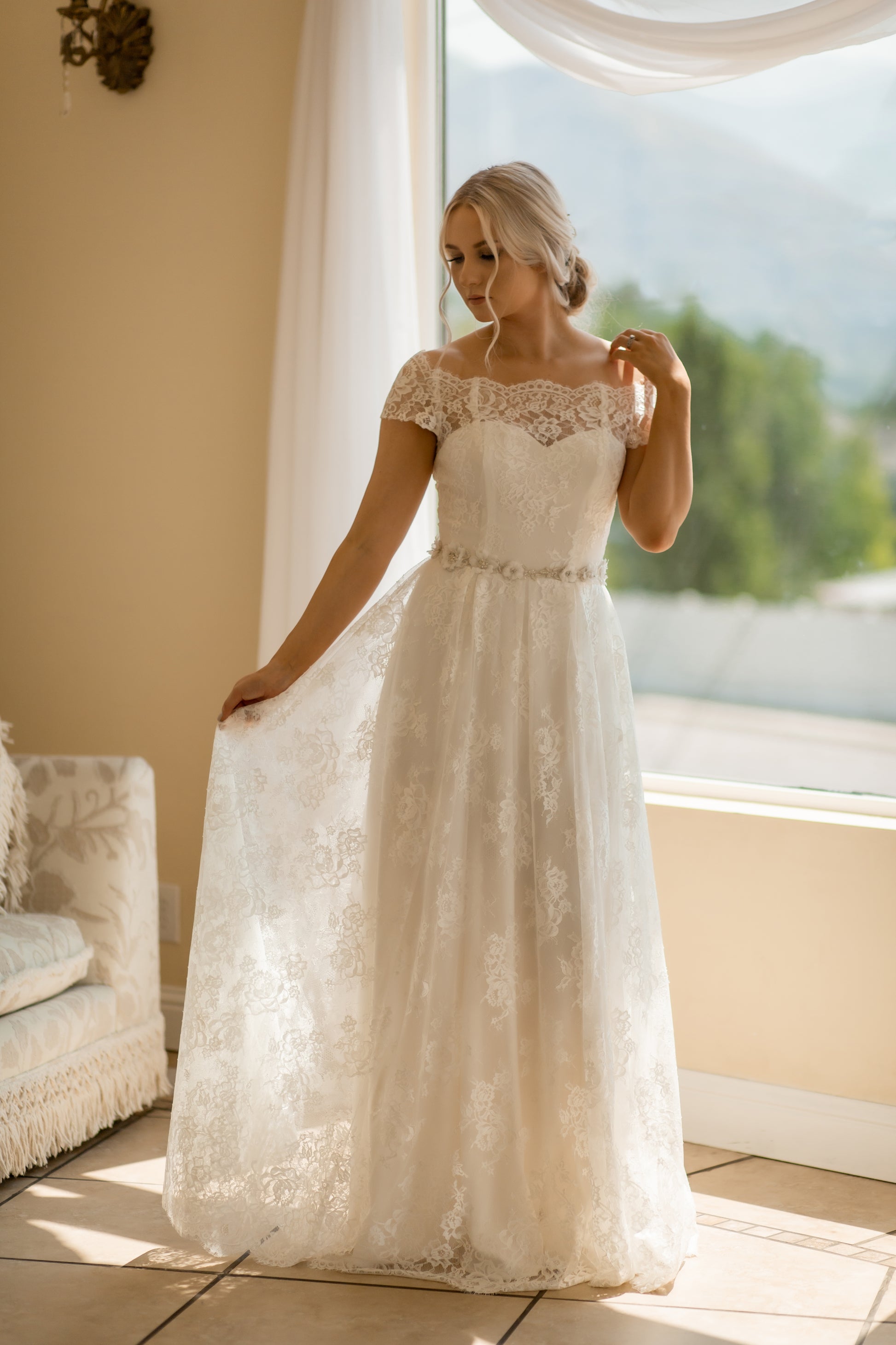 A full front image of the Serafina gown on a bride; she holds out the lace skirt showing off the folds created by the Inverted box pleats and A-line silhouette. The gown has been paired wih the Heidi belt.