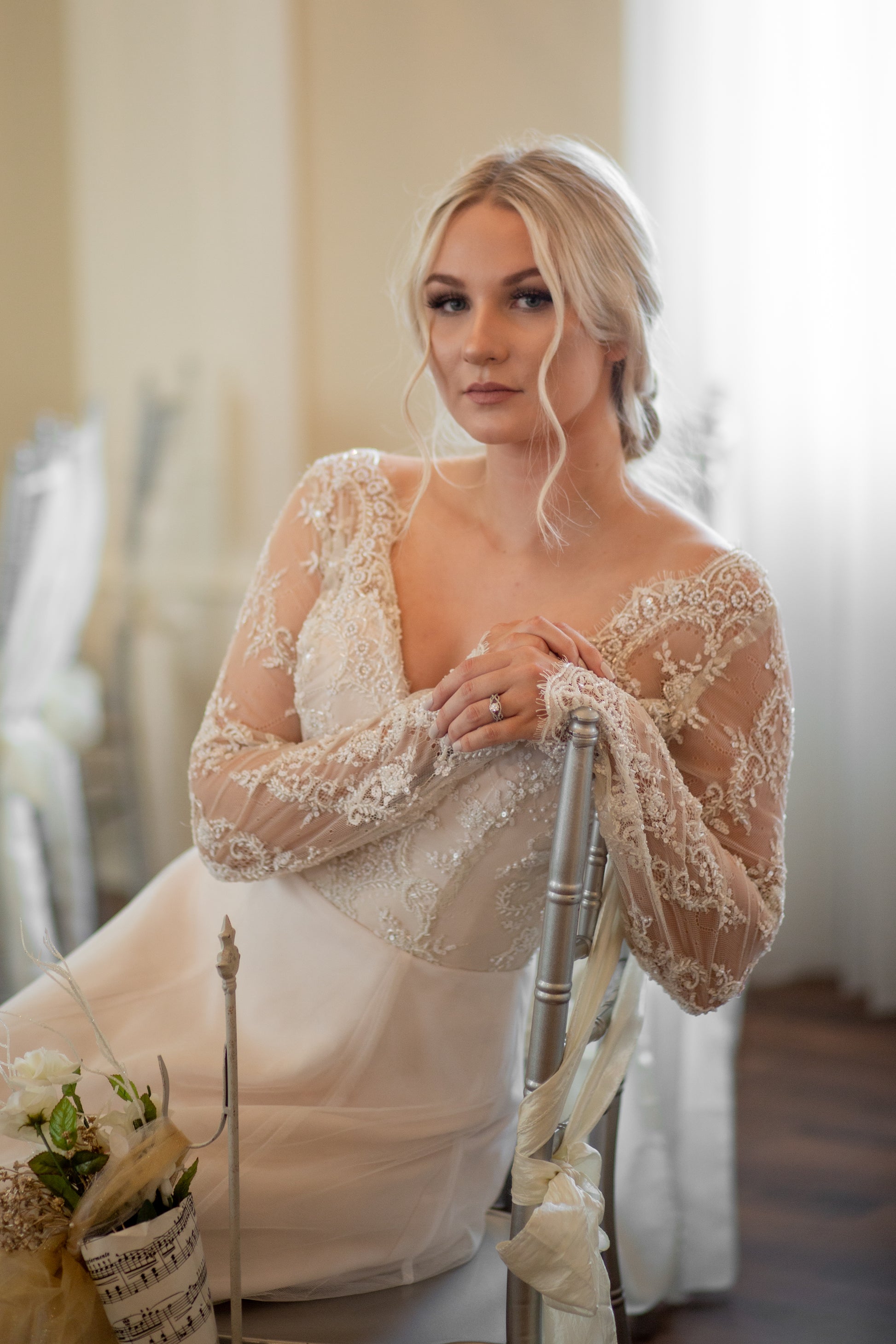Bride sits sideways on a chair and leans  with her arm over the back of the chair. The sleeves and lace covered bodice are shown off, along with the tiny waist created with a defined seam  .