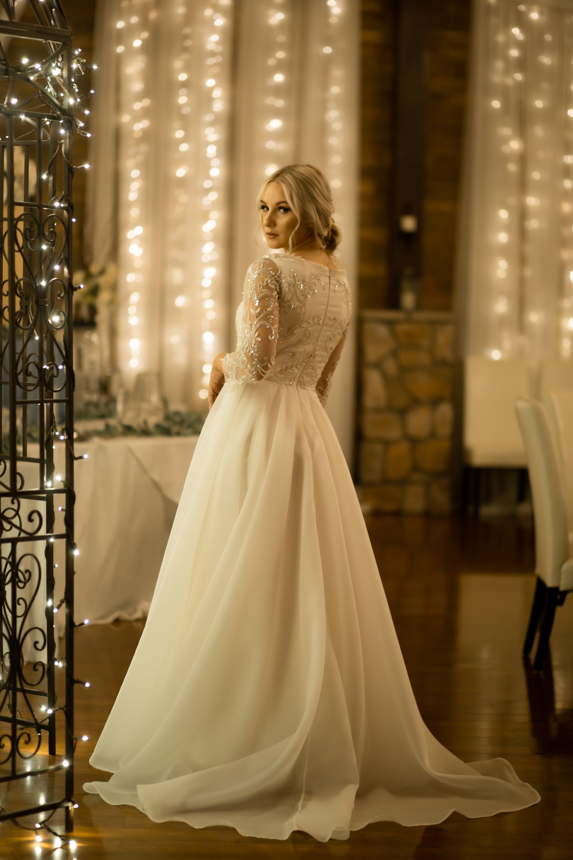 A styled shoot of a bride wearing the Theordora gown from the back, she leans to the side a little and looks over her shoulder. The gowns full skirt flows out while the organza train extends behind her.