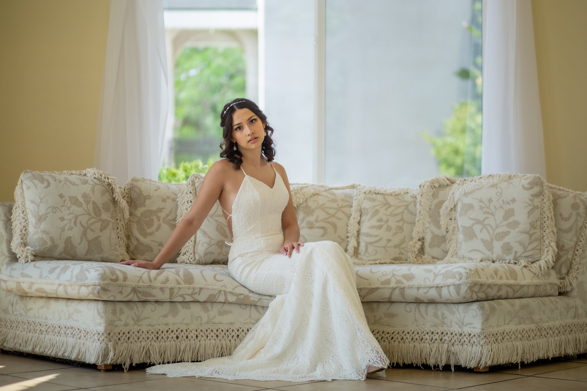 A bride sitting on a sofa in the Willoughby gown, she sits on the edge while leaning back on her hand. The train elegantly flares out to the side, while you can see the side cut and begining of the lace up detail.