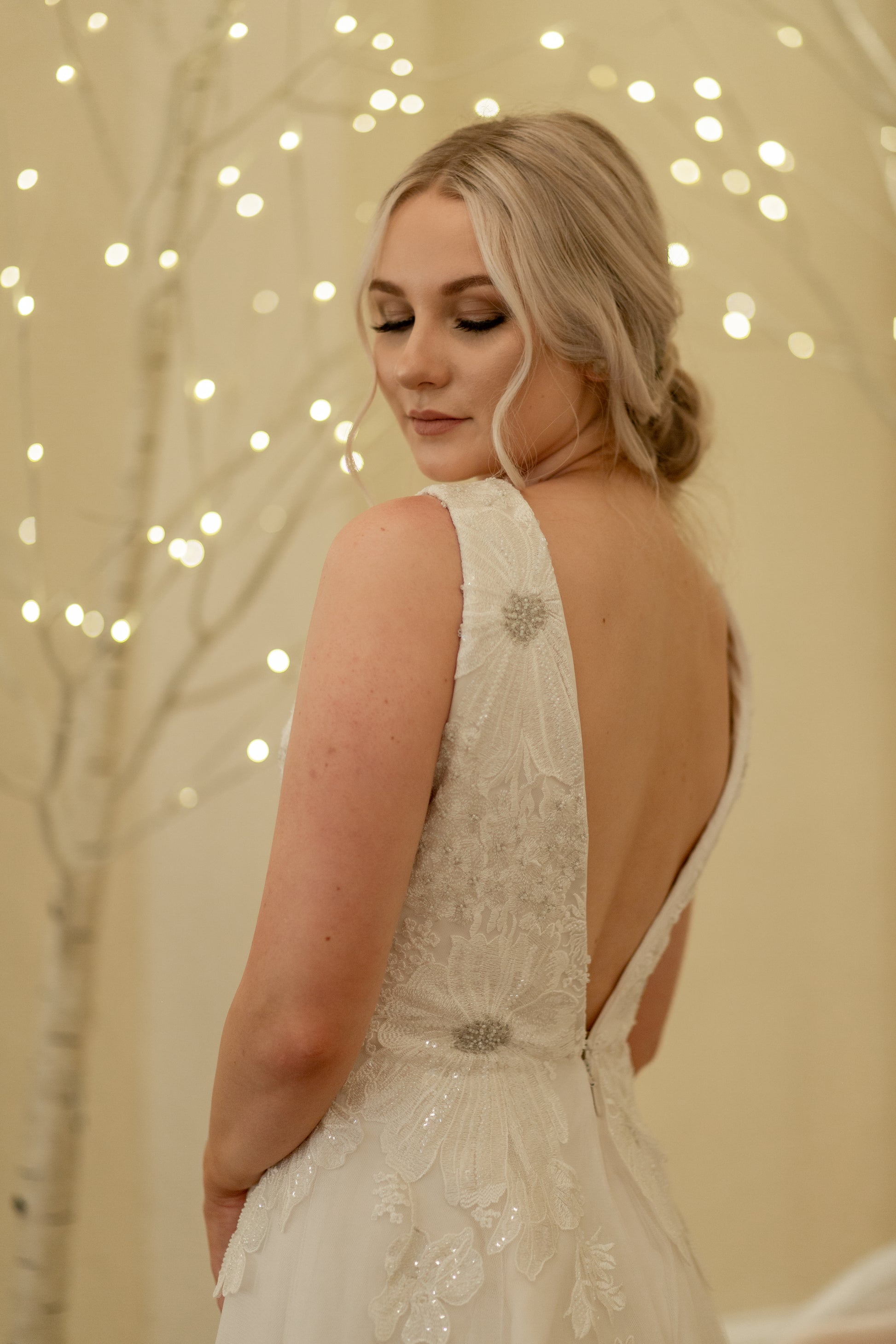 A bride wears the Poppy gown and looks back over her shoulder showing the back bodice, Beaded floral lace with metallic threading overlaying a structured satin bodice.