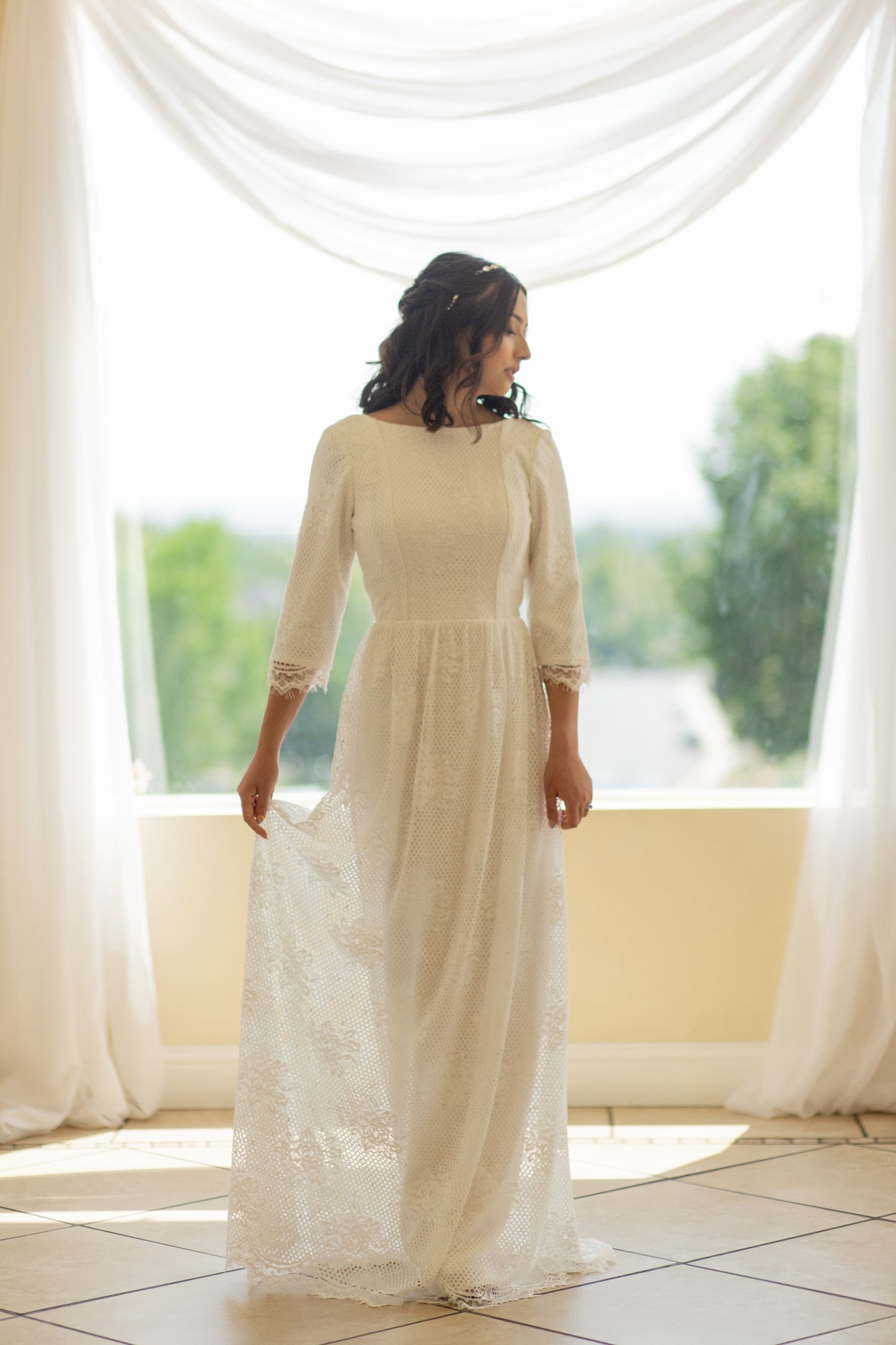 Bride wearing the Avabelle gown stands gracefully, showcasing the fitted bodice with wide boat neckline and three-quarter-length lace sleeves. The modified A-line silhouette flows gently into a soft lace skirt.