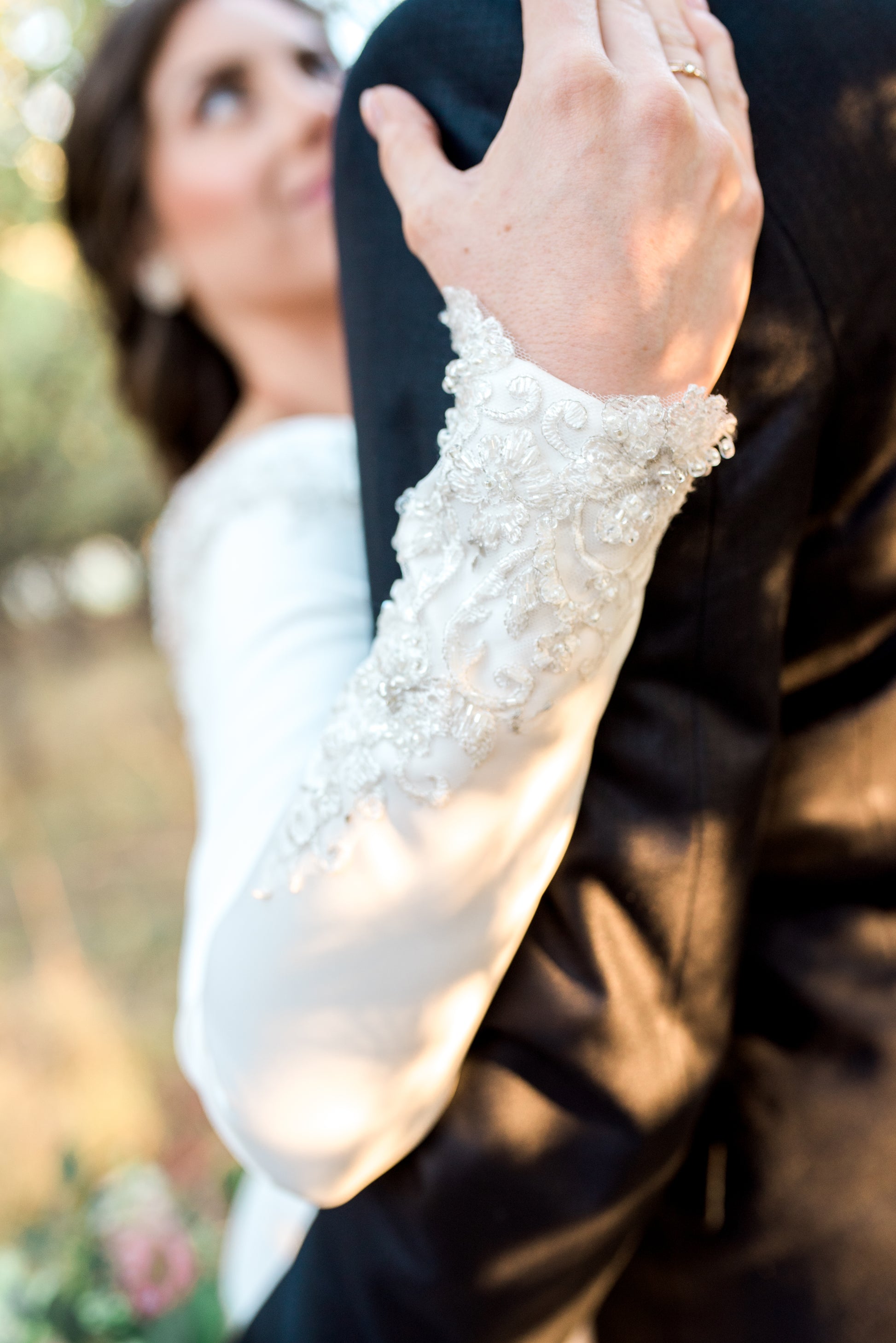 Detail shot of Ava gown's long satin sleeve ending in a delicate lace, the bride that wears it has her hand on the grooms shoulder.