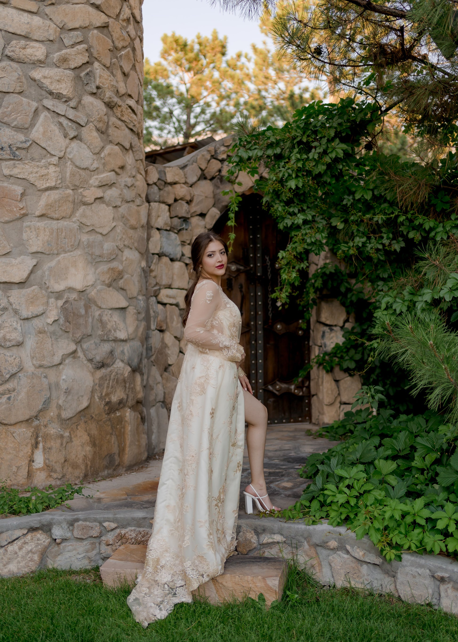 A full length back view of the Reagan gown on a bride as she steps up stone stairs. She twists back and looks at the camera with her leg showing through the wrap skirt. The train and lace sleeves are the focus.