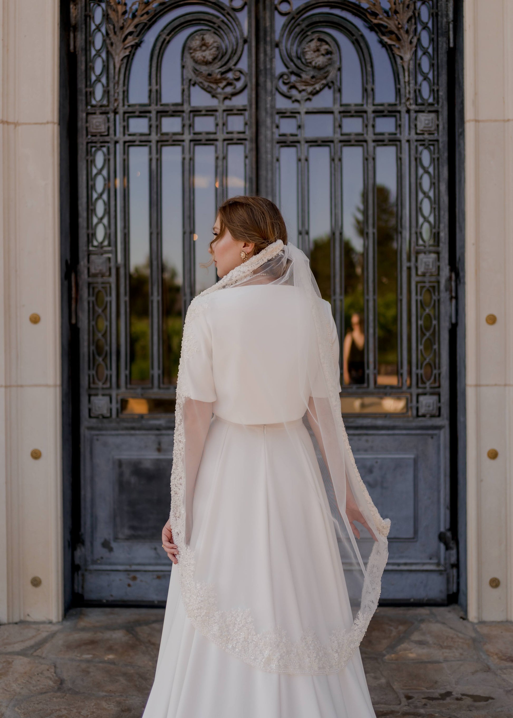 A photo of a bride from the back wearing the cassie set and ireland veil. She holds out the veil to the sides and faces away.