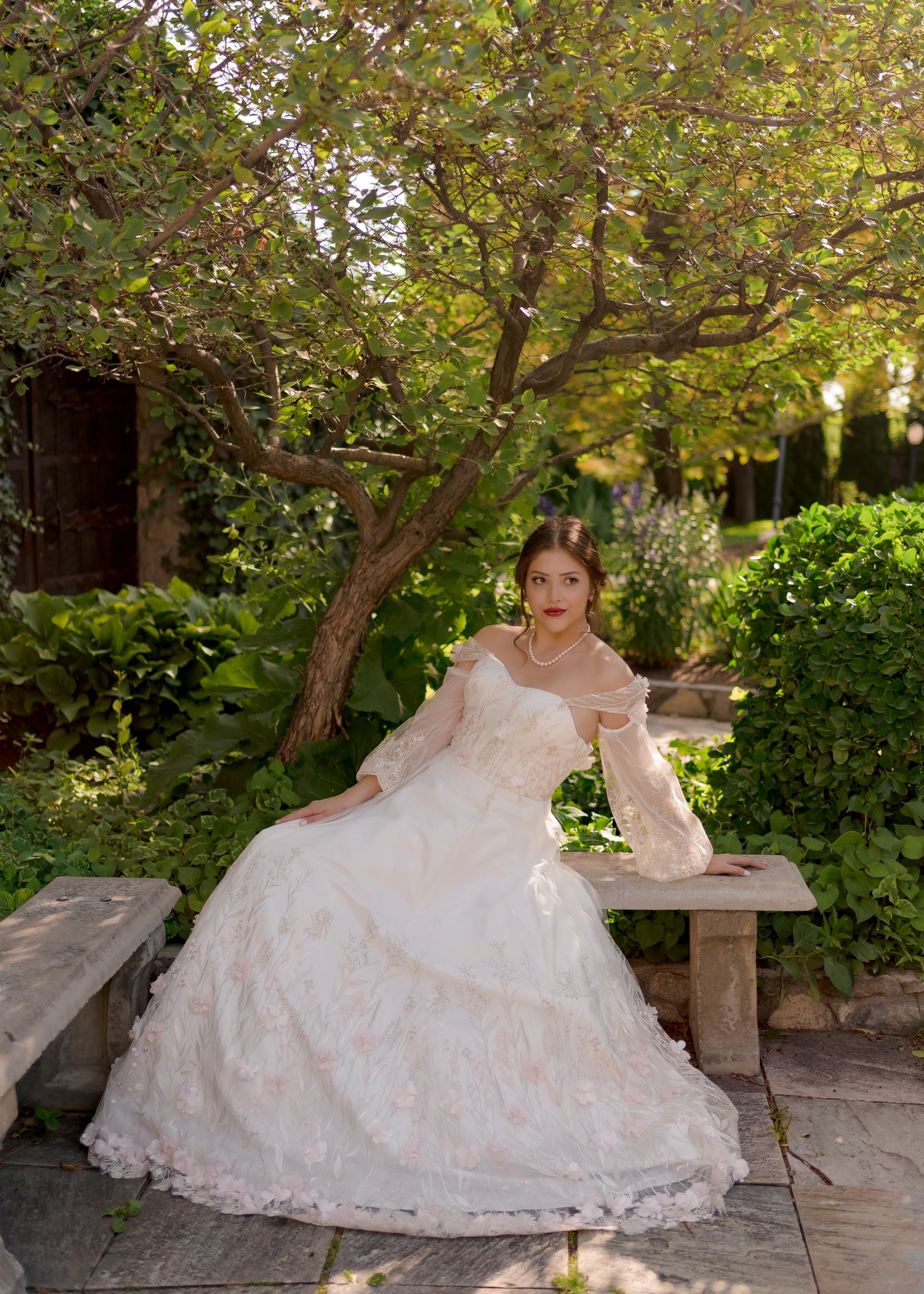 A photo of the Prue gown on a Bride; she sits on a bench in a garden. The Prue wedding gown has an a-line skirt, sweetheart neckline, and off the shoulder bishop sleeves.