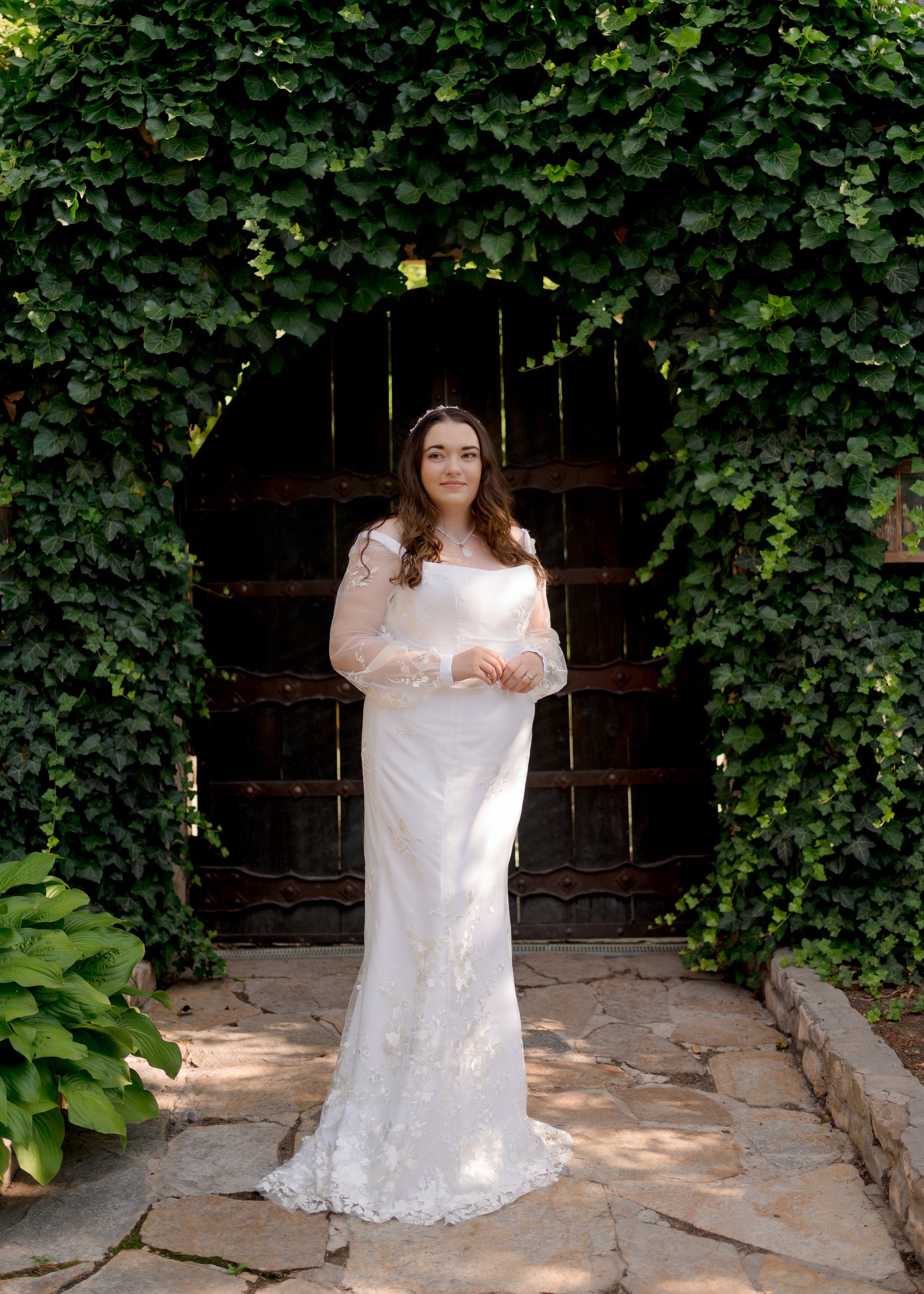 The violet gown on a model, she stands with her hands together in front of a wall of ivy; the gowns sleeves and trumpet shape are highlighted.
