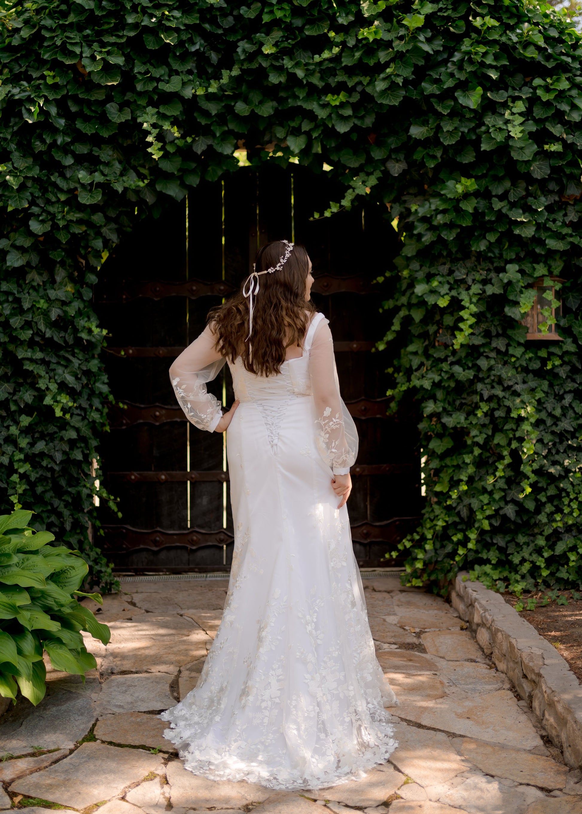 A full length back shot of the Violet wedding gown on a bride in front of a wall covered in ivy. The bridal gown's silhouette, structured bodice, lace up back, and small train are all featured.