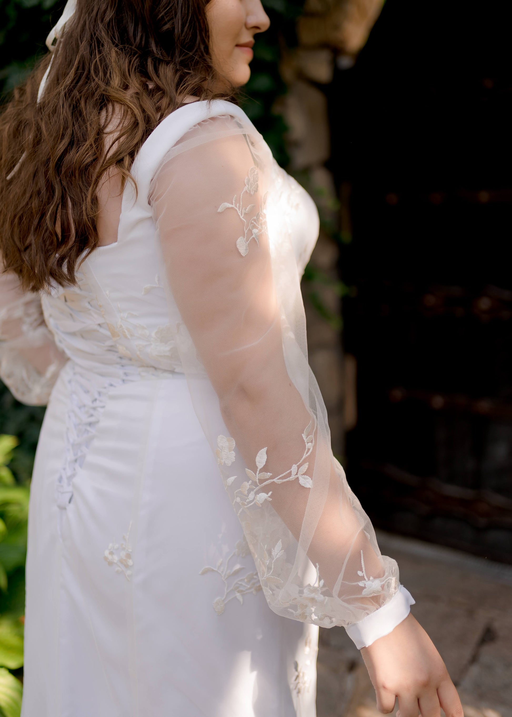 An angled back shot of a bride in the Violet gown; this shot is focused on the lace placement on the bishop sleeves and top skirt. The lace has smaller motifs at the top of the bodice, skirt, and sleeve and the motifs get larger the further down they get.