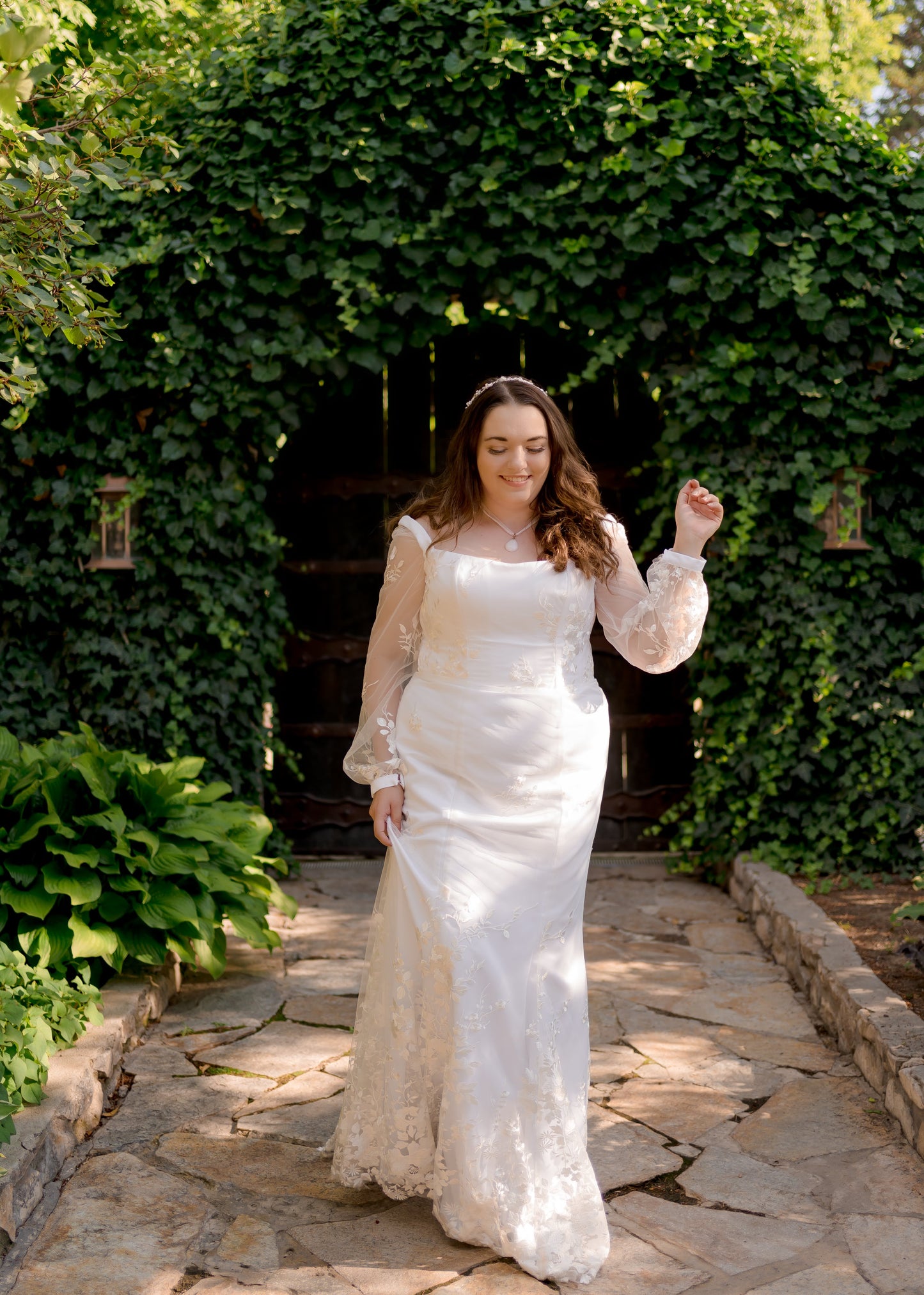 A walking shot of a bride wearing the Violet gown, she holds the skirt up with one hand while the gowns bishop sleeves, square neckline, and structured bodice are all in focus.
