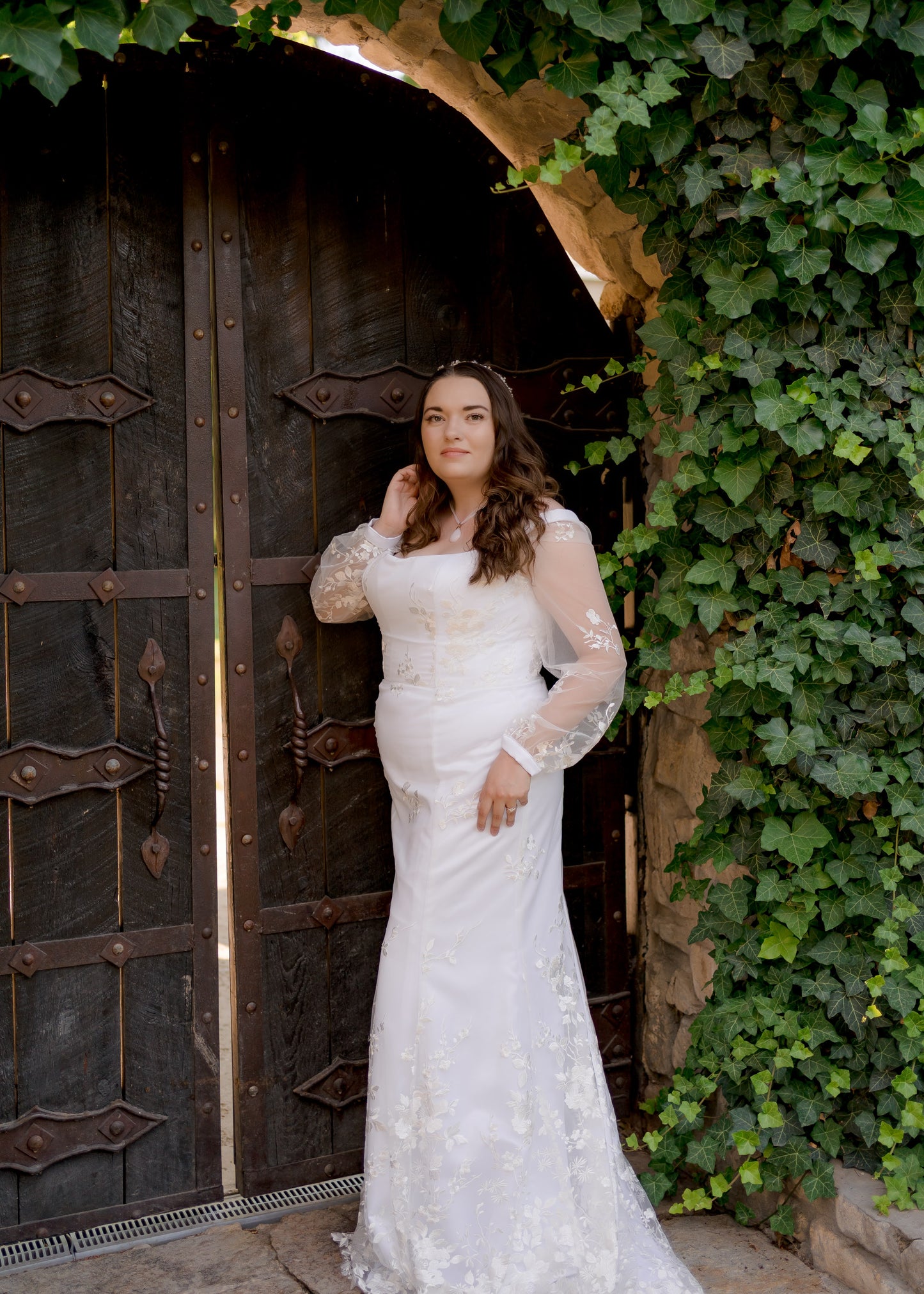 A full length shot of the violet wedding gown in front of a wooden door; notice the fit and flare silhouette, Sheer bishop lace sleeves with a cuff and the corseted fit.