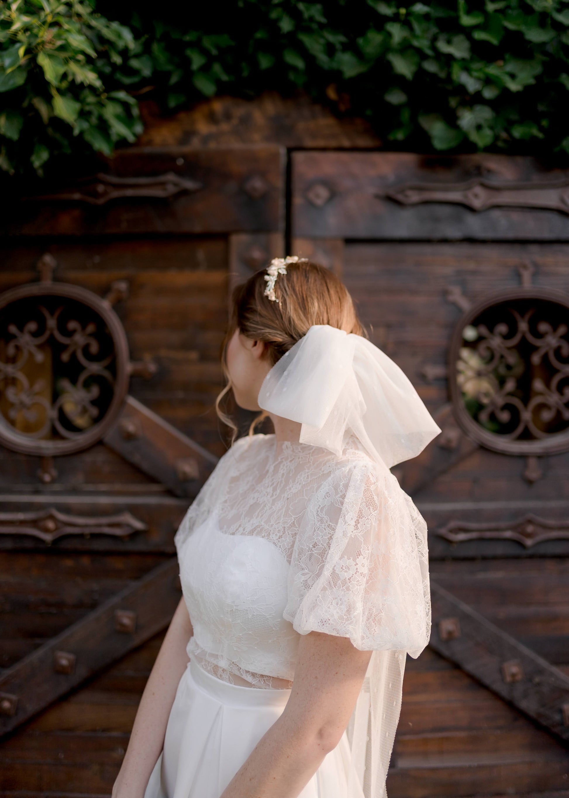 A bride wears the bridal bow veil, tess, in a swiss dot fabric long tails. The bride is wearing our jessa gowm and is looking away showing off the bow portion of the veil as the tails hang hidden behind her back.