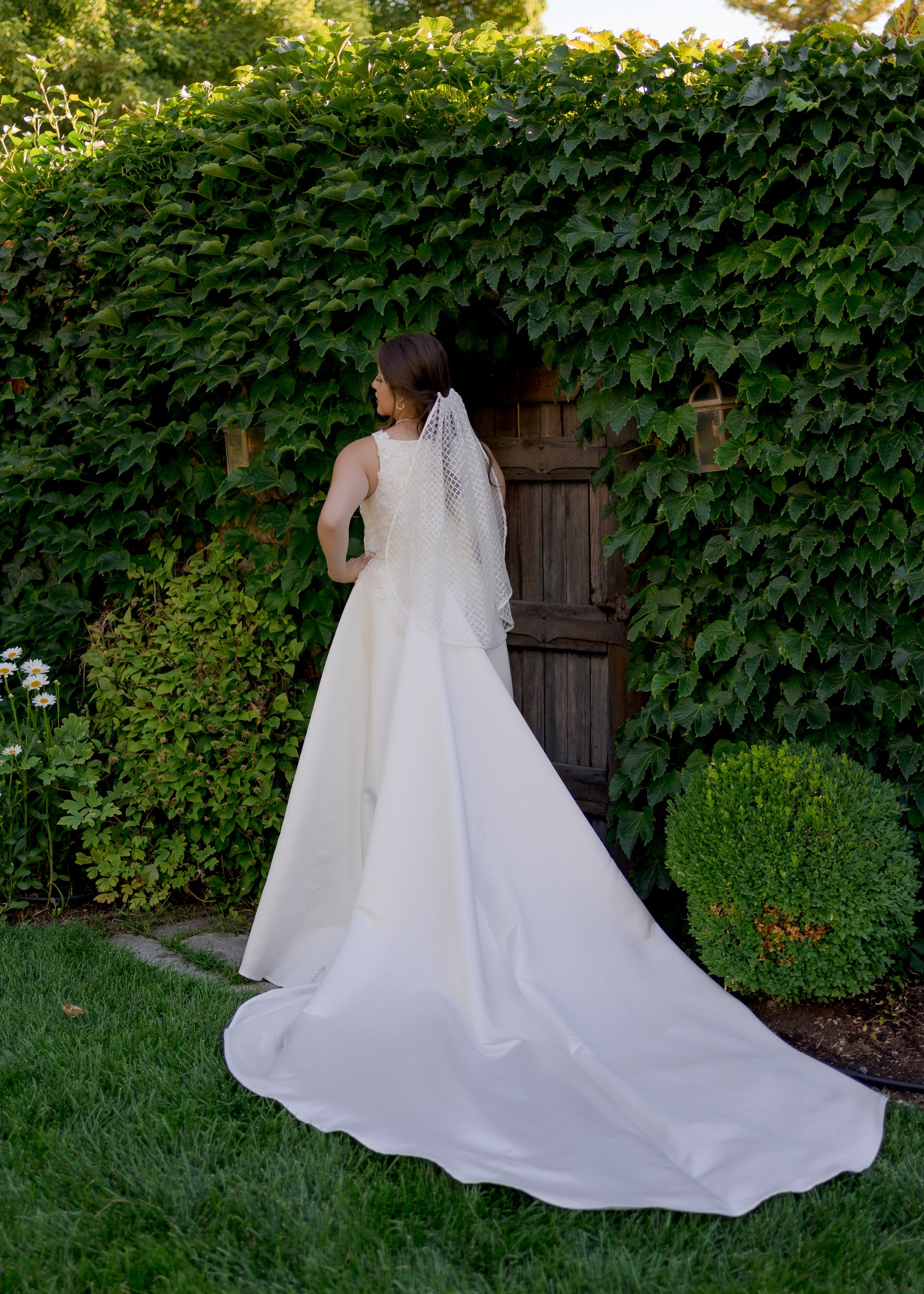 This photo is of a bride in front of an ivy covered wall with a Bride in the sparkly diamond tulle veil, Gloria, and our satin and lace gown, Sabina. The bride faces away from the camera so we can see the fingertip length veil in her hair and the extra long tain extending out on the grass.