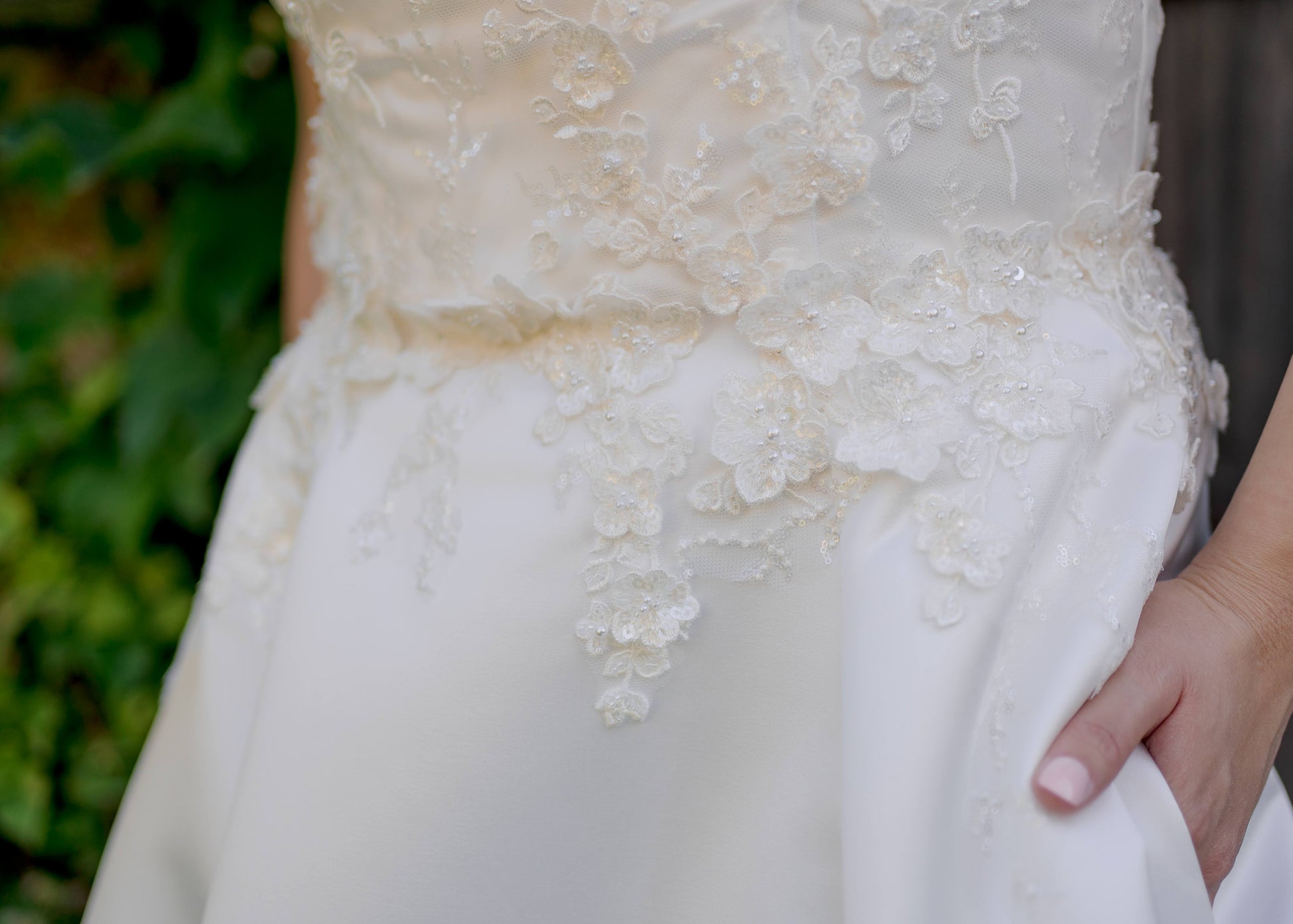 A close up of the Sabina gowns's waist; the lace tapers from the bodice into the folds of the satin skirt. The bride wearing the gown also has her hand in her pocket at the side seam  .