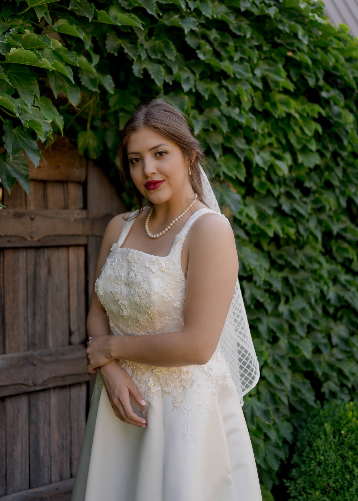 A photo of one of our brides wearing the sabina gown, 3d lace bodice and satin skirt, paired with the gloria veil, Ribbon-edged bridal veil with shimmer sequins in a diamond pattern. The fingertip length veil just pokes out frm behind her shoulder.