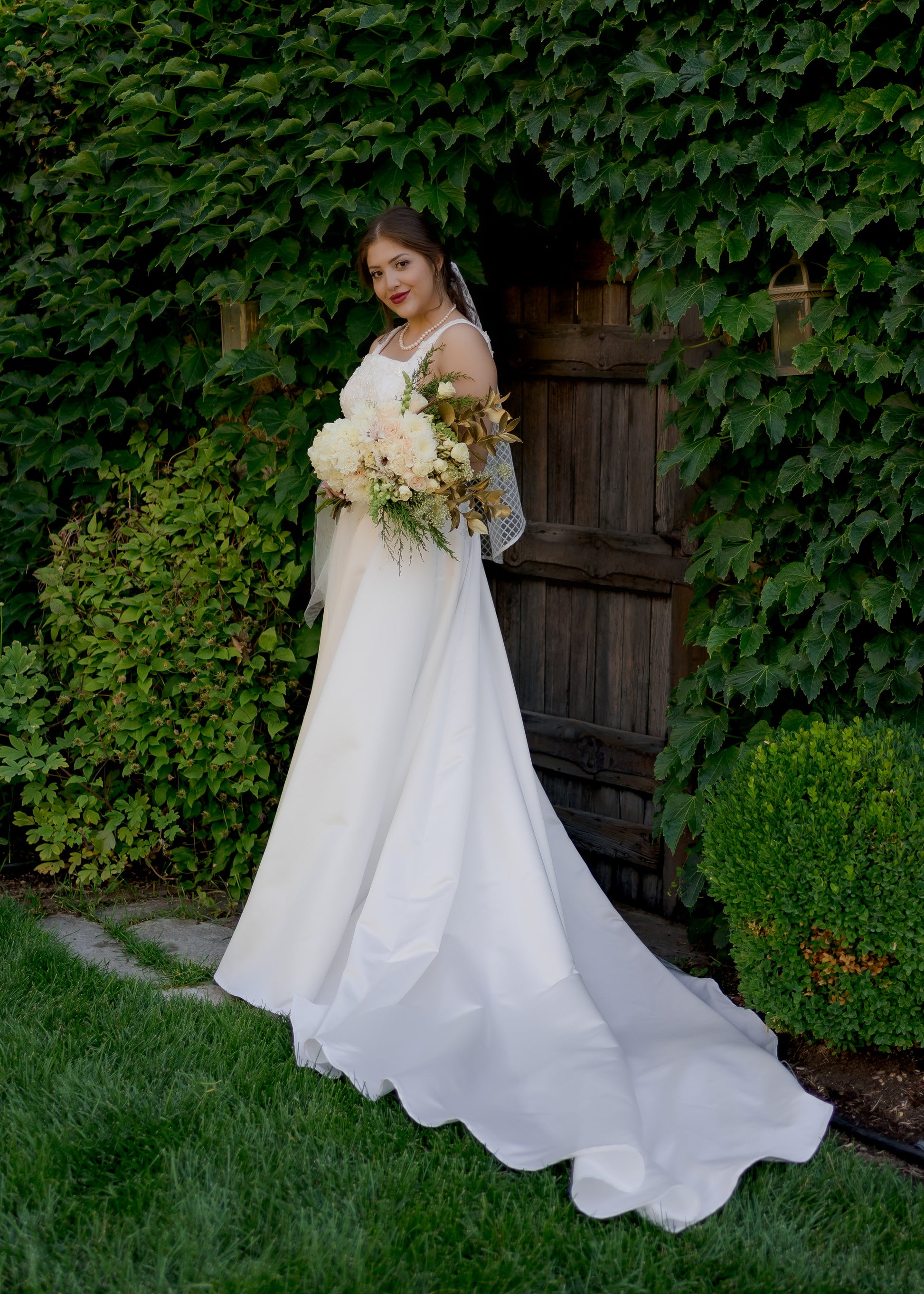 A side view of the Sabina bridal gown on a bride; the satin train is extended to the side of her while she looks toward the camera. The regal gown is paired with a fingertip length Gloria veil.