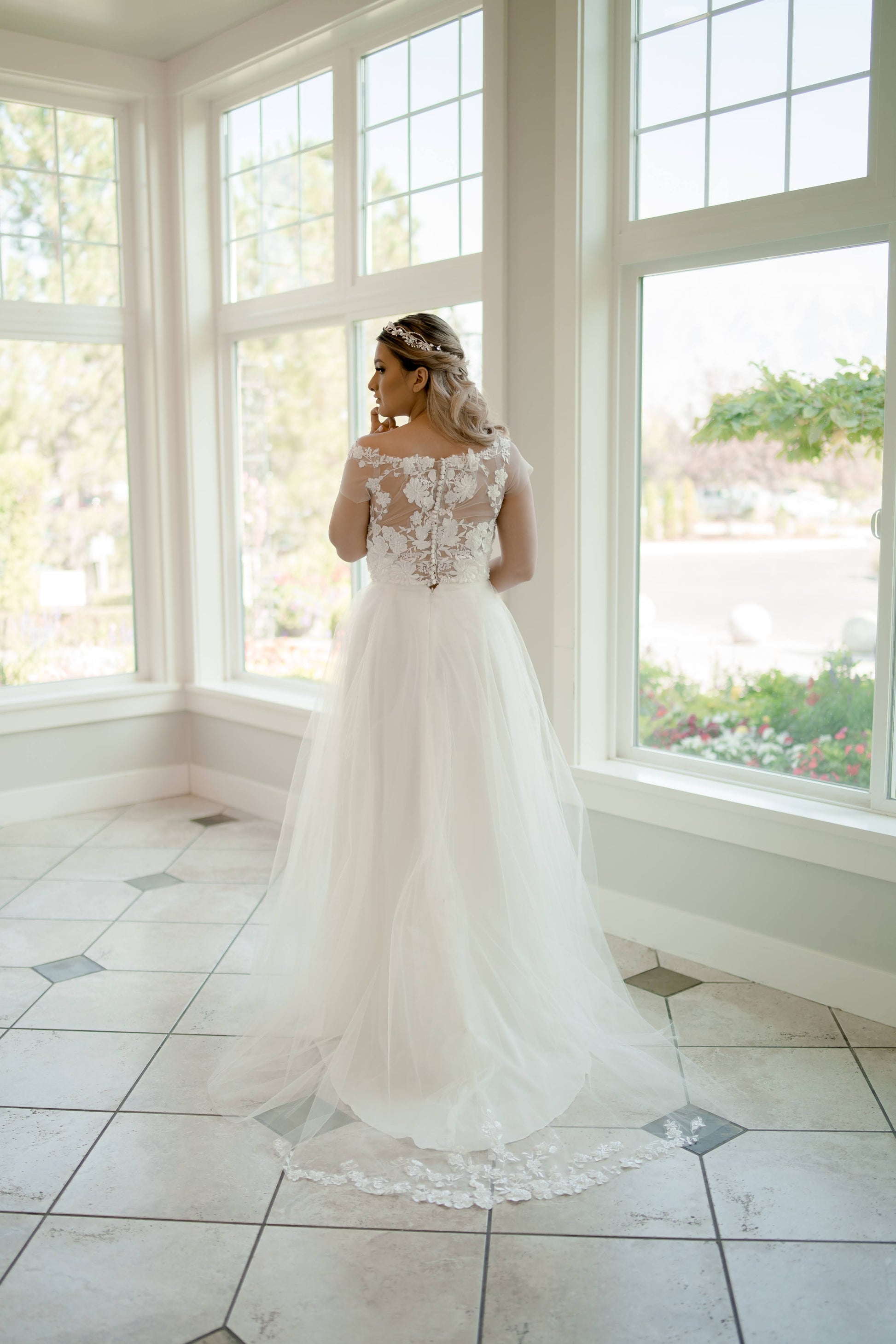 Full-length back shot of the model turned away, showing both the satin and tulle trains with lace detail at the hem
