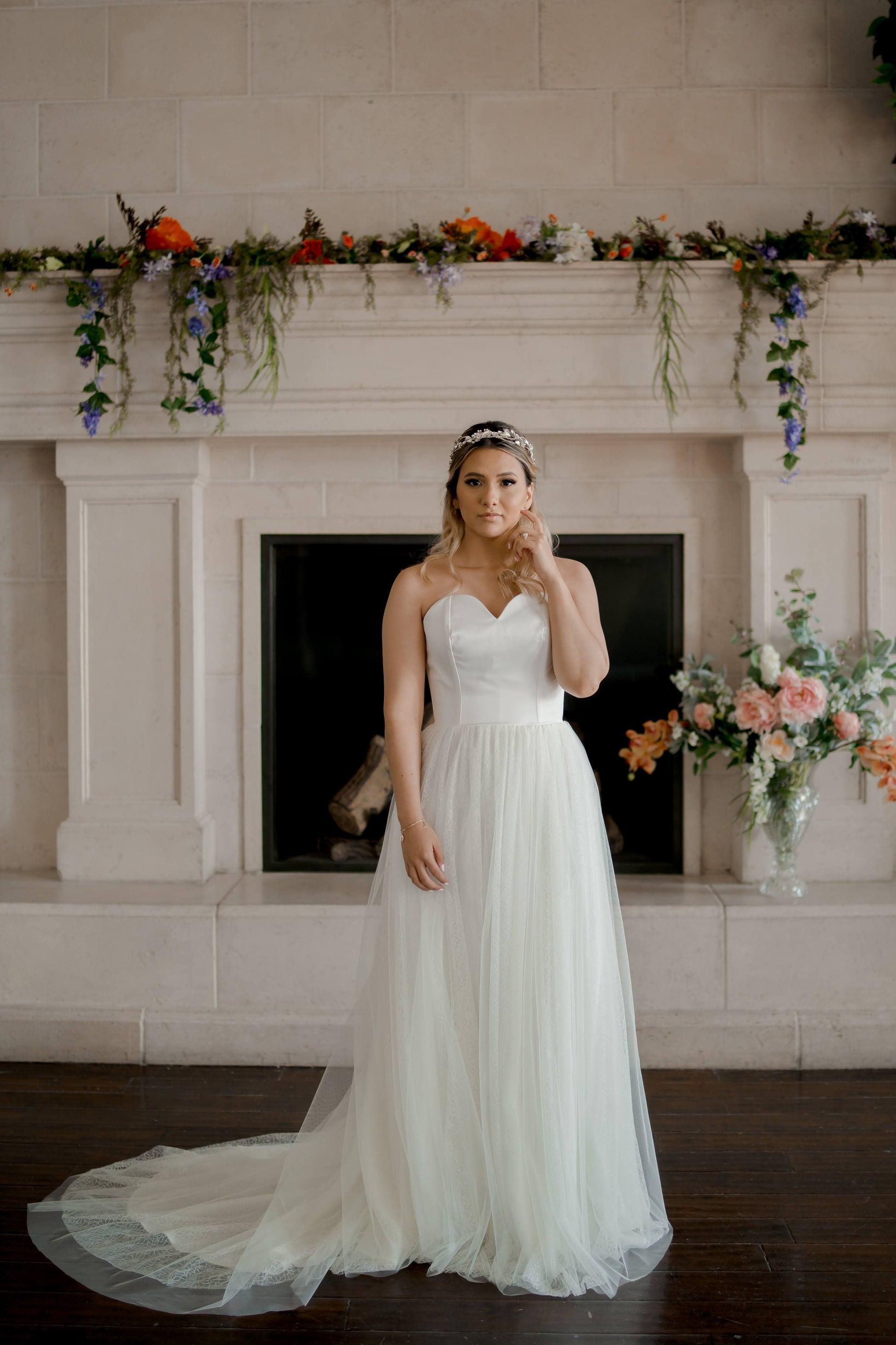 Bride wearing the Beatrice A-line wedding gown stands in front of a fireplace. The soft layers of tulle and daisy lace gently cascade to the floor, with the romantic sweetheart neckline and subtle train creating an elegant, timeless bridal look.